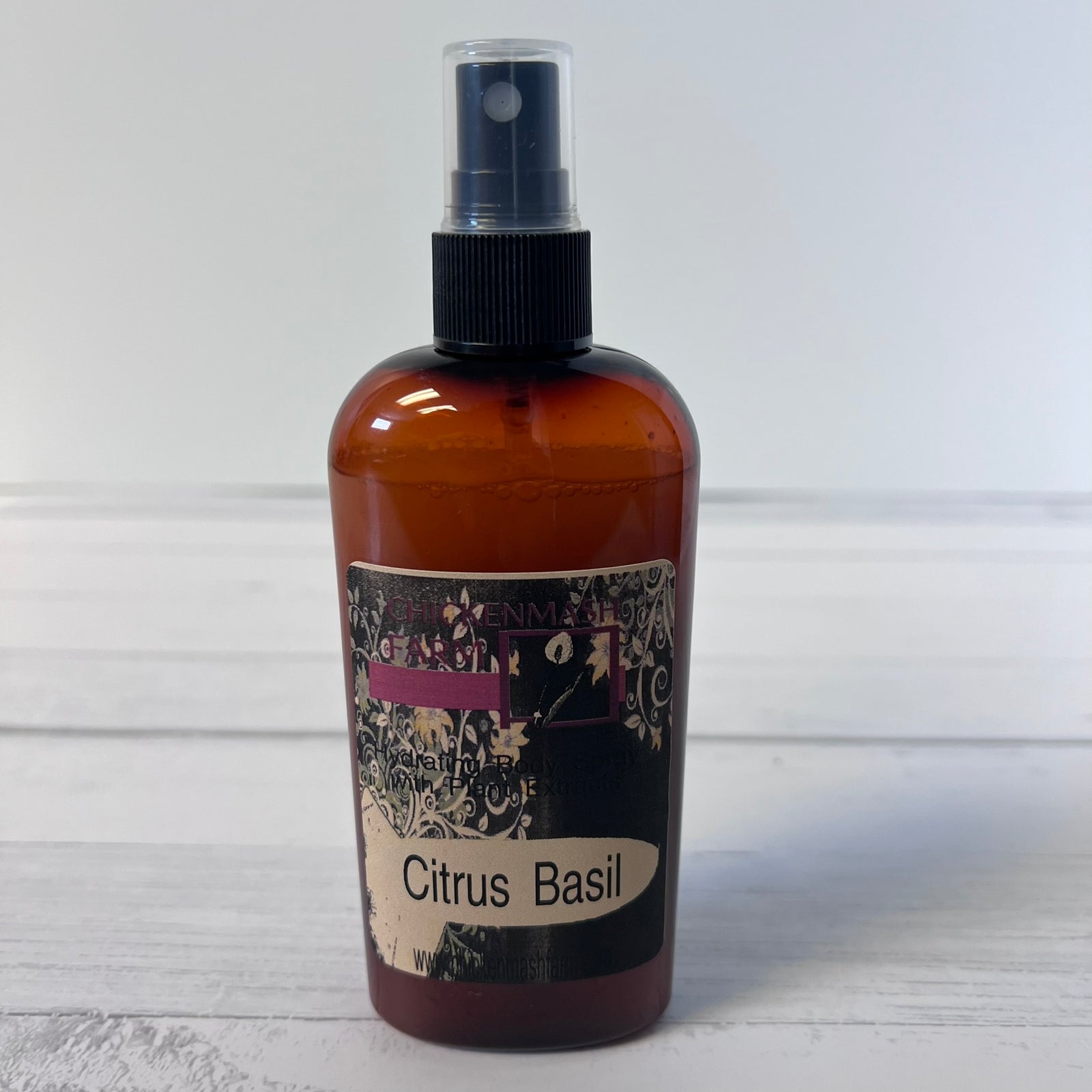 An amber spray bottle with a black spray top and a decorative label reading 'Citrus Basil' on a white wood background.