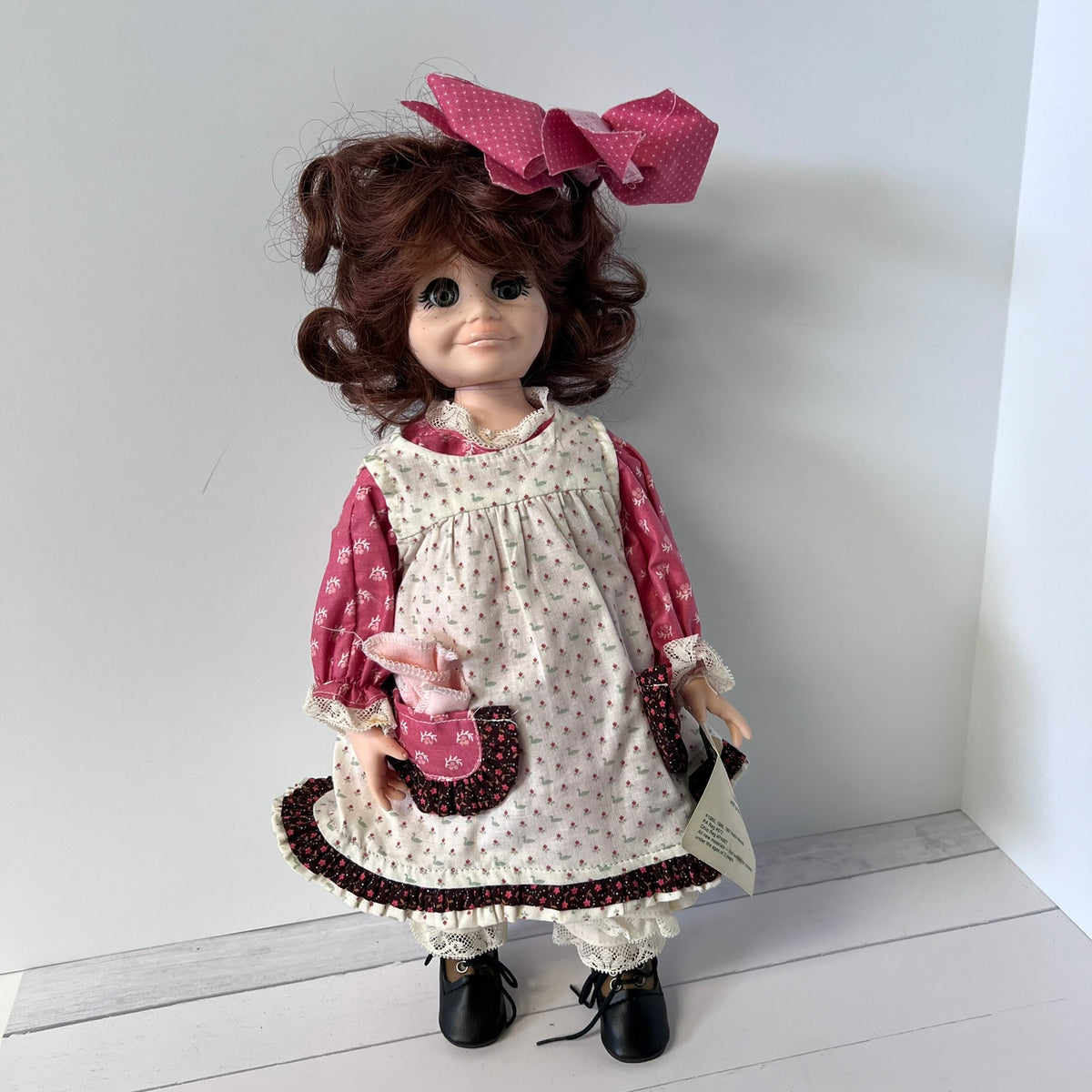 A vintage-style doll with curly brown hair, wearing a cream dress with pink floral patterns, a pink polka-dot apron, and a large pink bow, standing against a white wall.