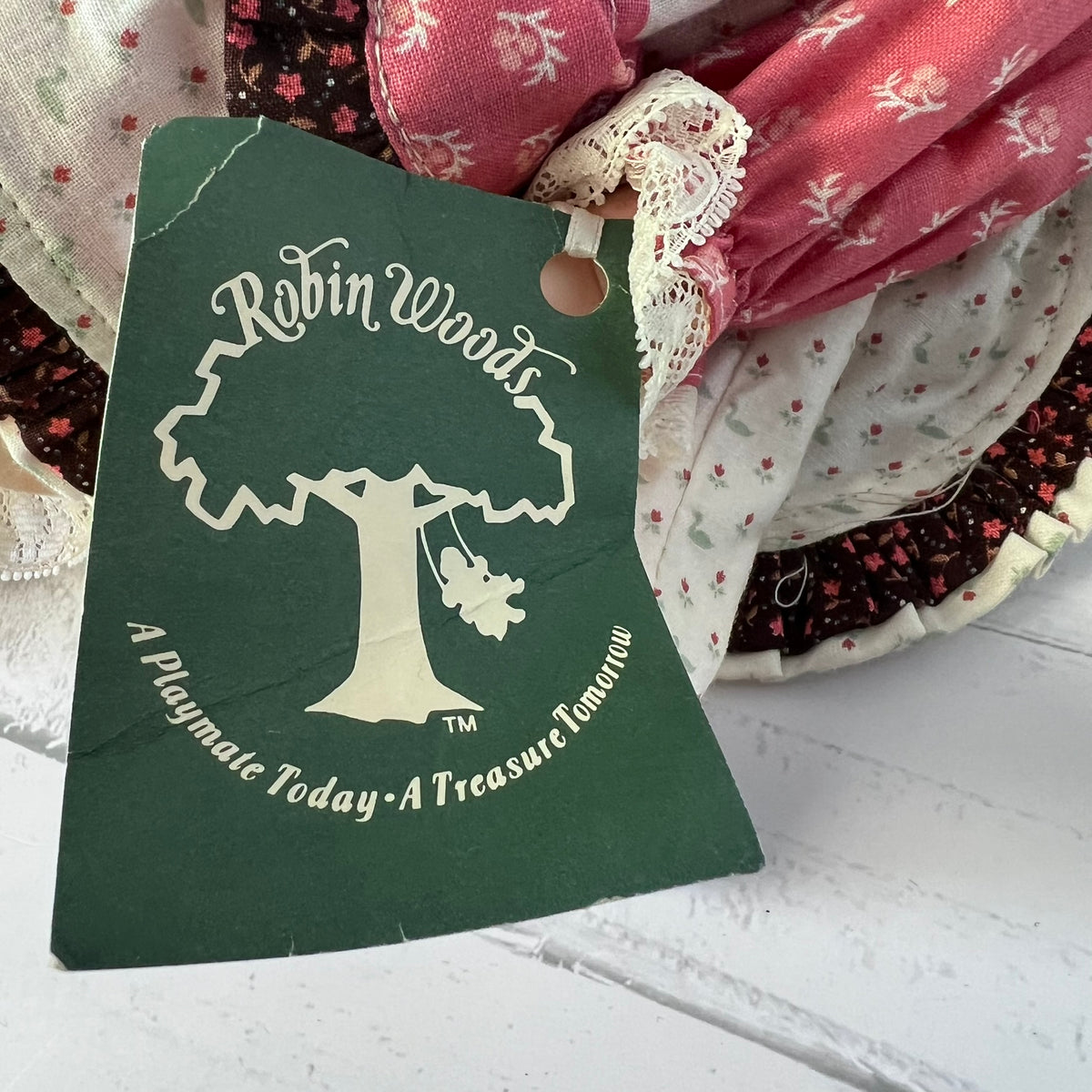 A dark green fabric tag with a white tree logo and the text 'Robin Woods' and 'A Playmate Today - A Treasure Tomorrow' attached to a bundle of vintage patterned fabrics.