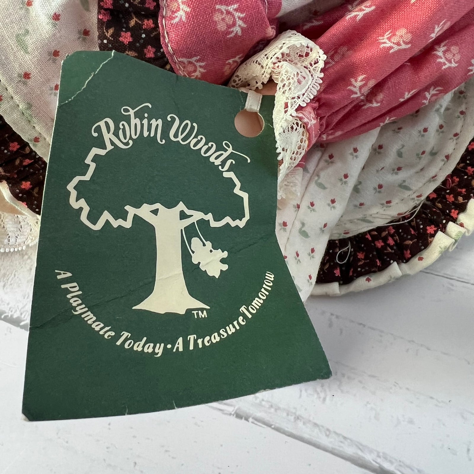 A dark green fabric tag with a white tree logo and the text 'Robin Woods' and 'A Playmate Today - A Treasure Tomorrow' attached to a bundle of vintage patterned fabrics.