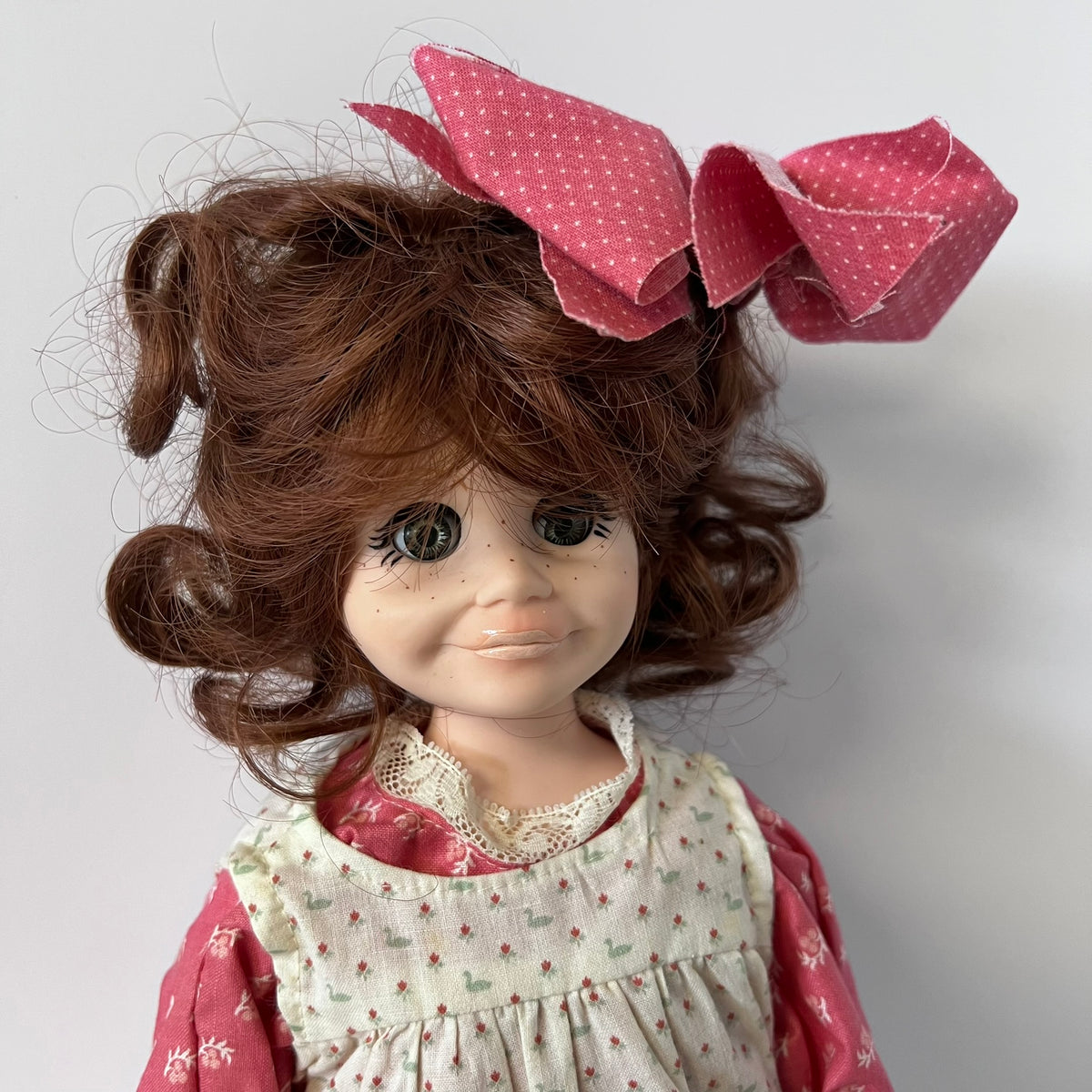A vintage-style doll with curly brown hair, a large pink polka-dot bow, and a cream dress with small red and green floral patterns, shown against a plain light gray background.