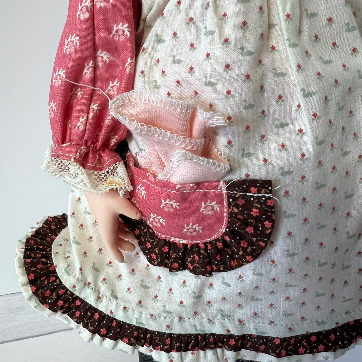 A close-up of a handmade doll or toy dress with a cream linen base, small red and green floral print, a pink patterned sleeve, and a matching floral apron-style front panel with lace trim and ruffles.