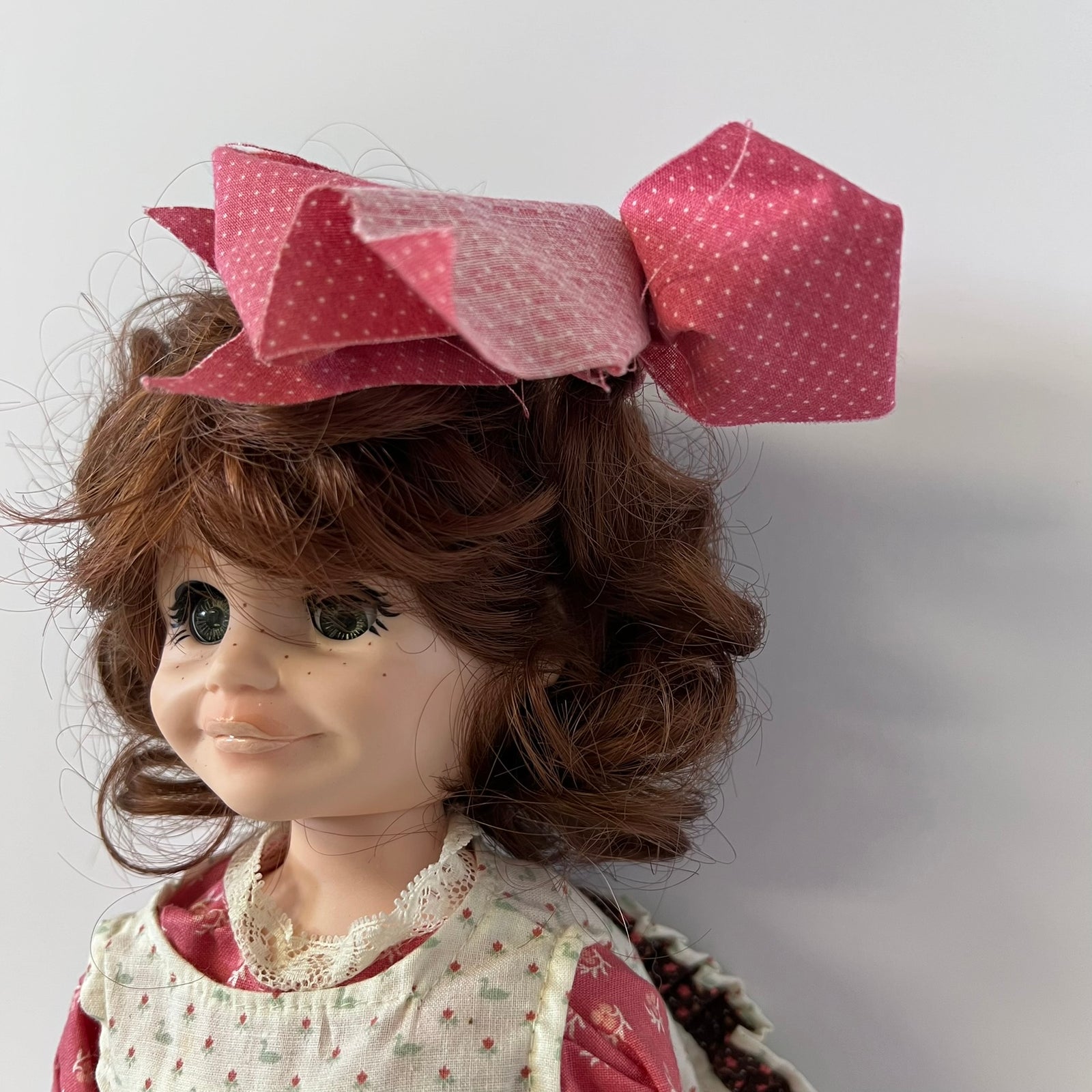 A close-up of a doll with brown curly hair wearing a large pink polka-dot bow headband and a cream dress with small red and green floral patterns.