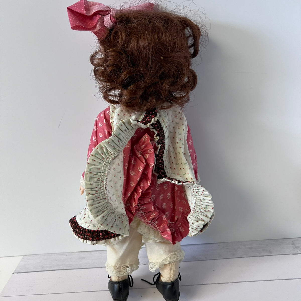 A vintage-style doll viewed from behind, featuring curly brown hair, a pink bow, and a ruffled pink and cream dress with white pants and black shoes, set against a plain light gray wall.