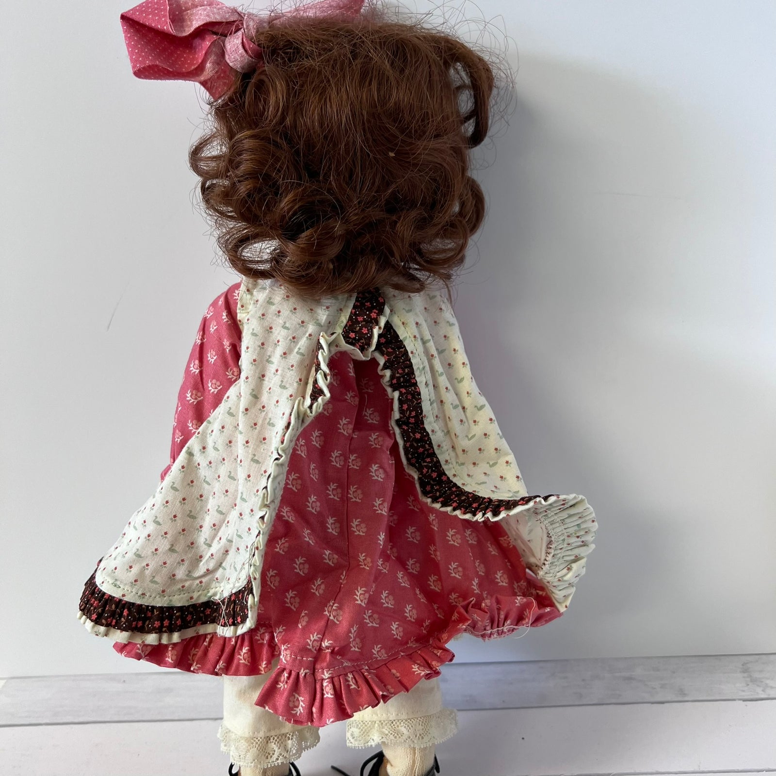 A vintage-style doll viewed from the back, featuring curly brown hair, a pink bow, and a multi-layered dress with a red patterned bodice, cream floral cape sleeves, and cream ruffled trim.
