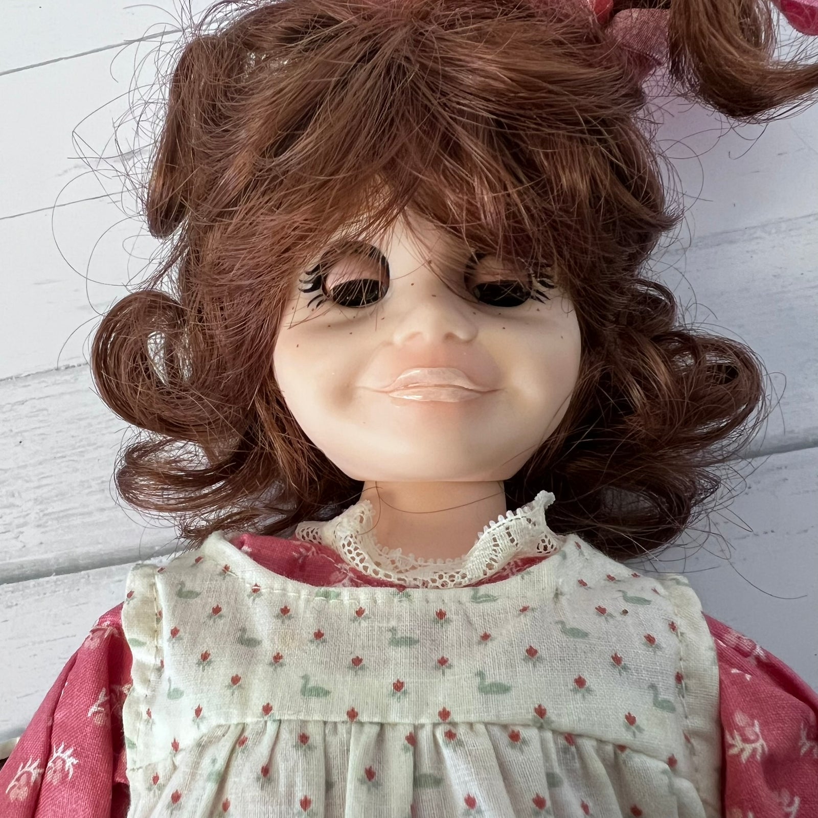 A close-up of a vintage-style doll with brown curly hair, a cream dress with a small floral pattern, and a pink long-sleeve underneath, set against a white wooden background.