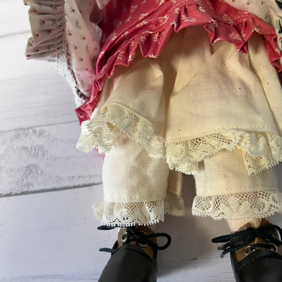 A close-up of a doll's lower body wearing a cream linen skirt with lace trim, a pink ruffled top, and black lace-up shoes, set against a white wooden background.