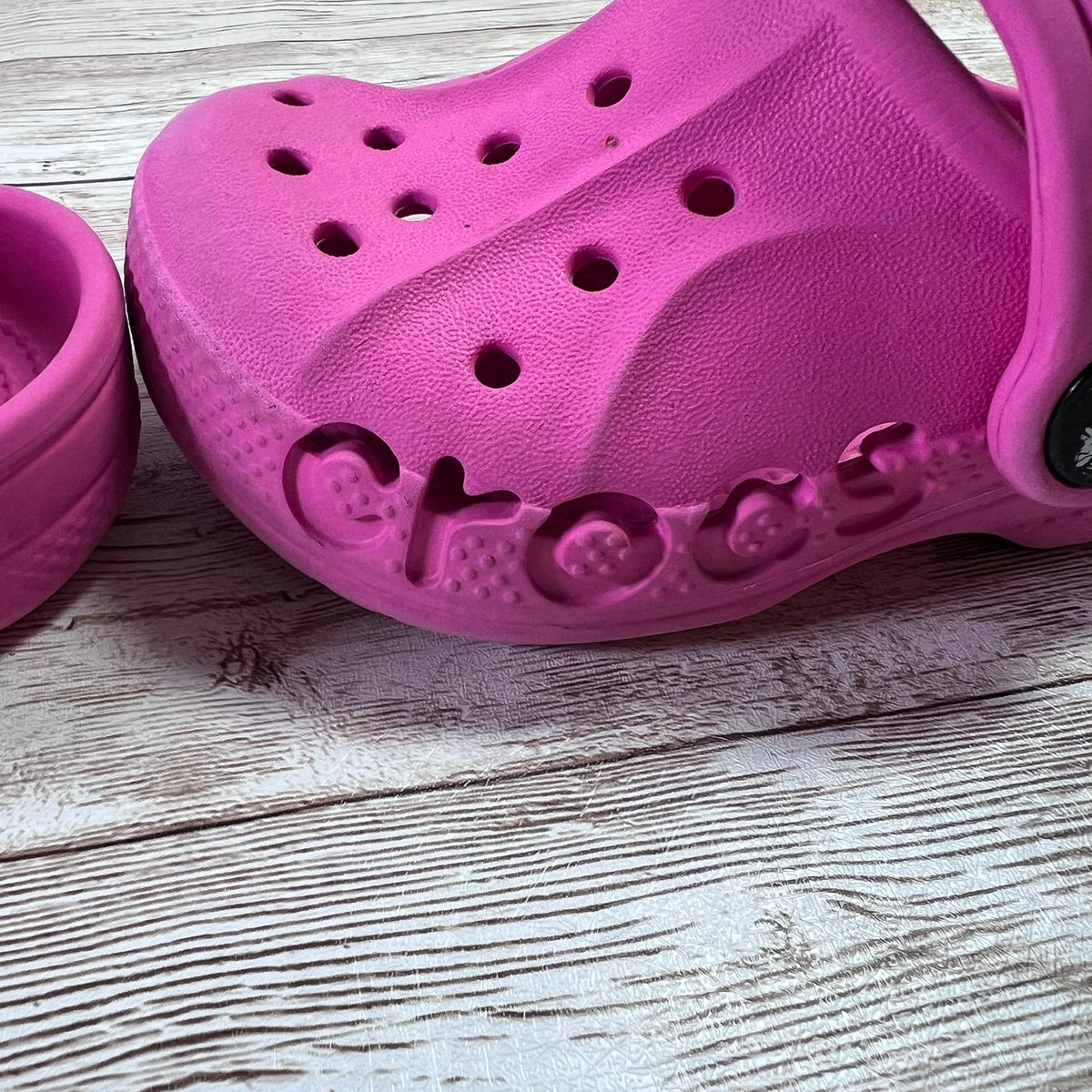 A bright pink Crocs clog shown on a light gray wood plank surface, with part of another matching pink Crocs shoe visible on the left.