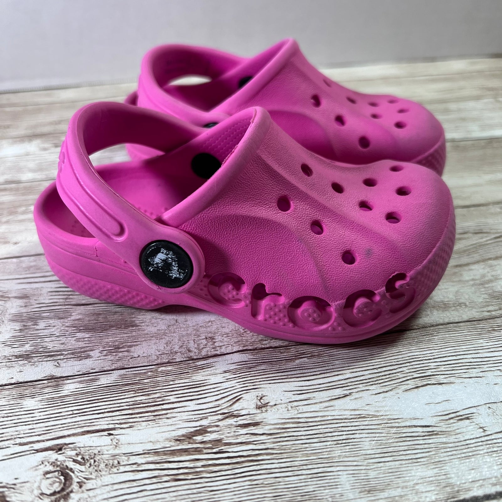 A pair of bright pink Croc-style clogs with vented uppers and a flexible sole, photographed on a light weathered wood surface.