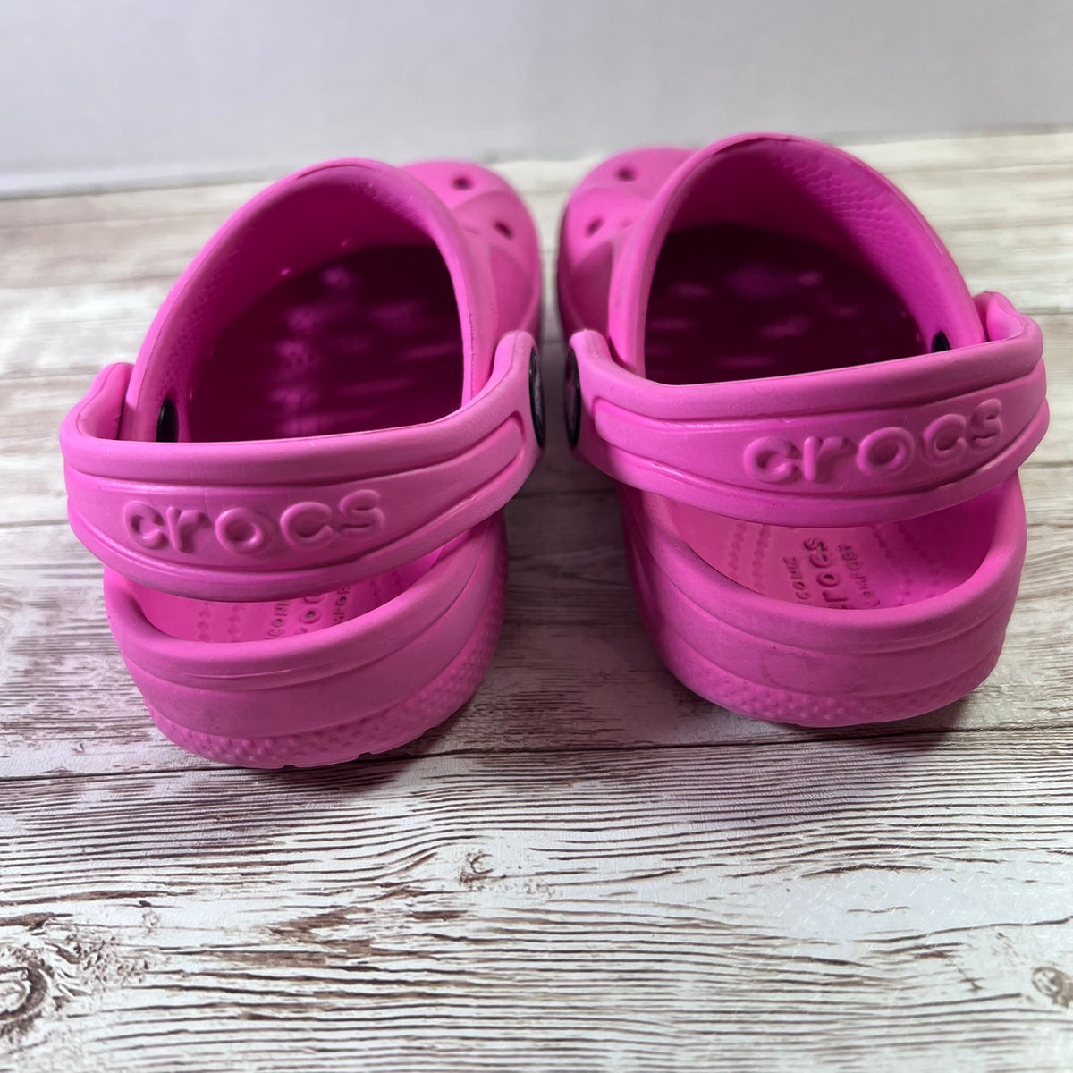 A pair of pink Crocs children's clogs shown from the back, resting on a light-colored wooden surface.