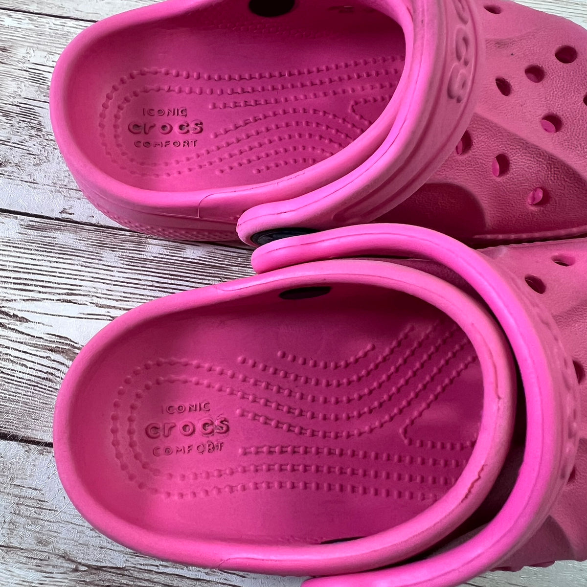 A pair of bright pink Crocs clogs shown from the top on a rustic whitewashed wood surface. The Crocs brand name and 'ICE CLOG COMFORT' text are visible on the soles.
