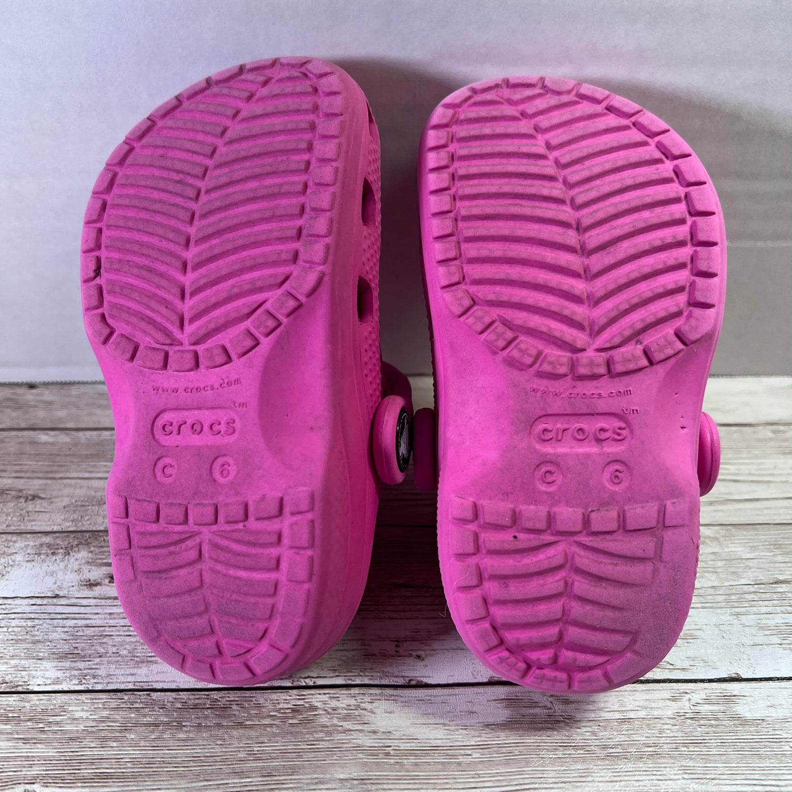 A pair of pink Crocs clogs shown from the sole side, resting on a light gray wooden surface against a light gray background.