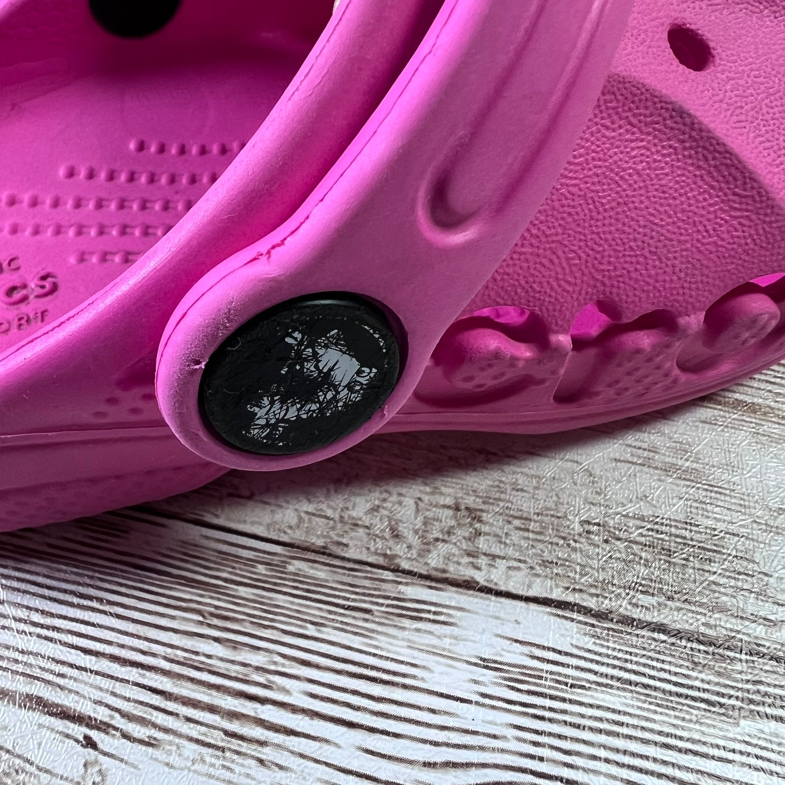 A close-up of a bright pink Croc-style clog shoe resting on a light-colored wood surface, showing the textured strap, footbed, and a black circular attachment point.