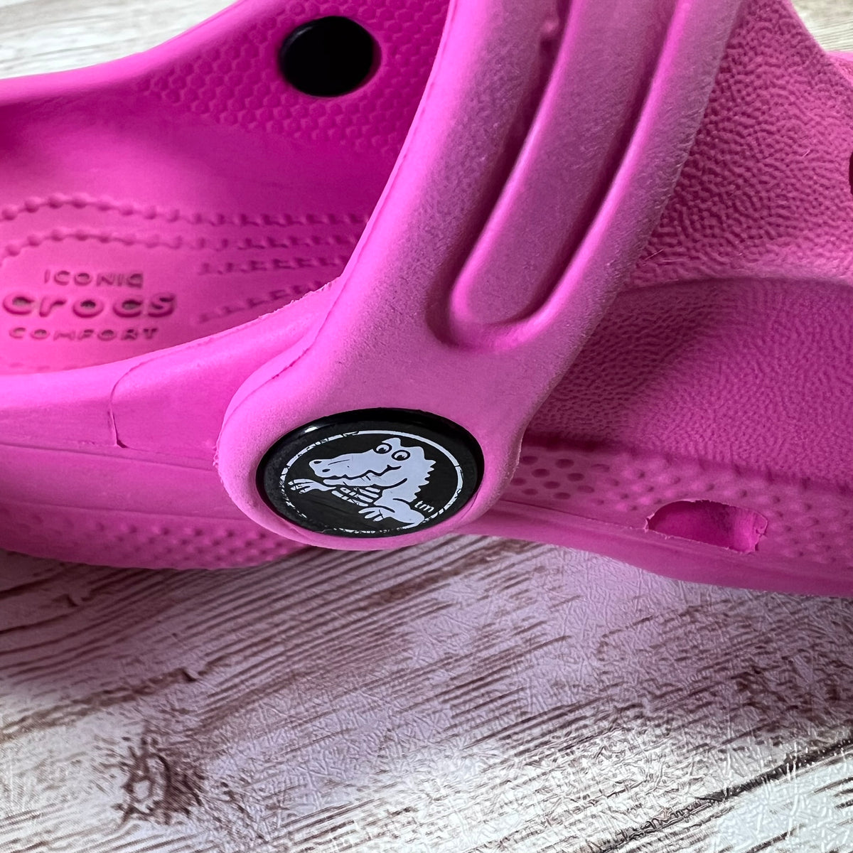 A bright pink Croc-style clog shoe photographed close-up on a light wood surface. The insole shows embossed Croc branding, and the side has a circular black patch with the Croc alligator logo.