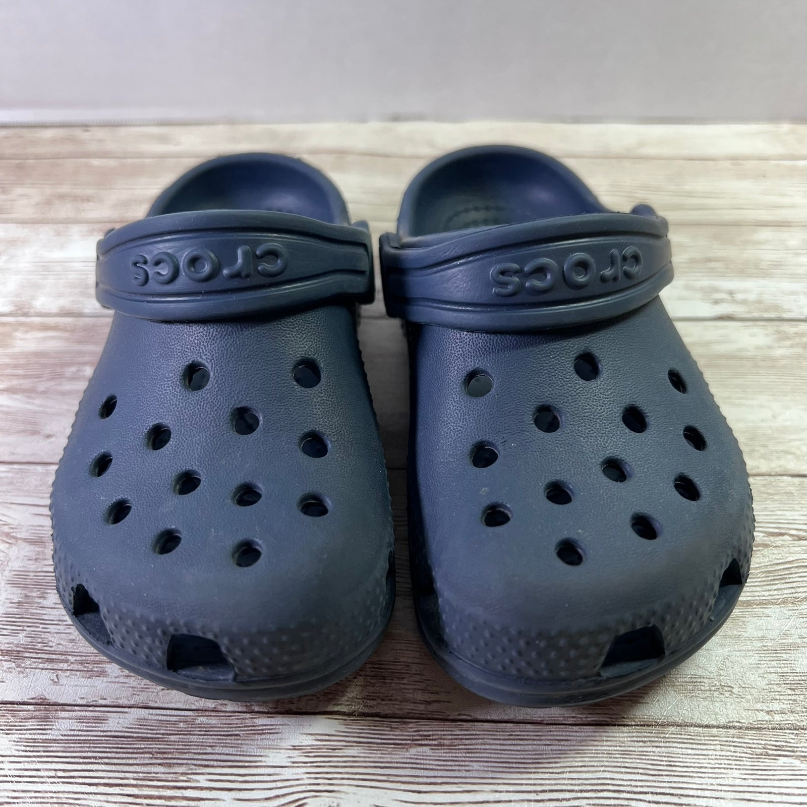 A pair of dark navy blue Crocs clogs placed on a light-colored wooden surface against a neutral gray wall.
