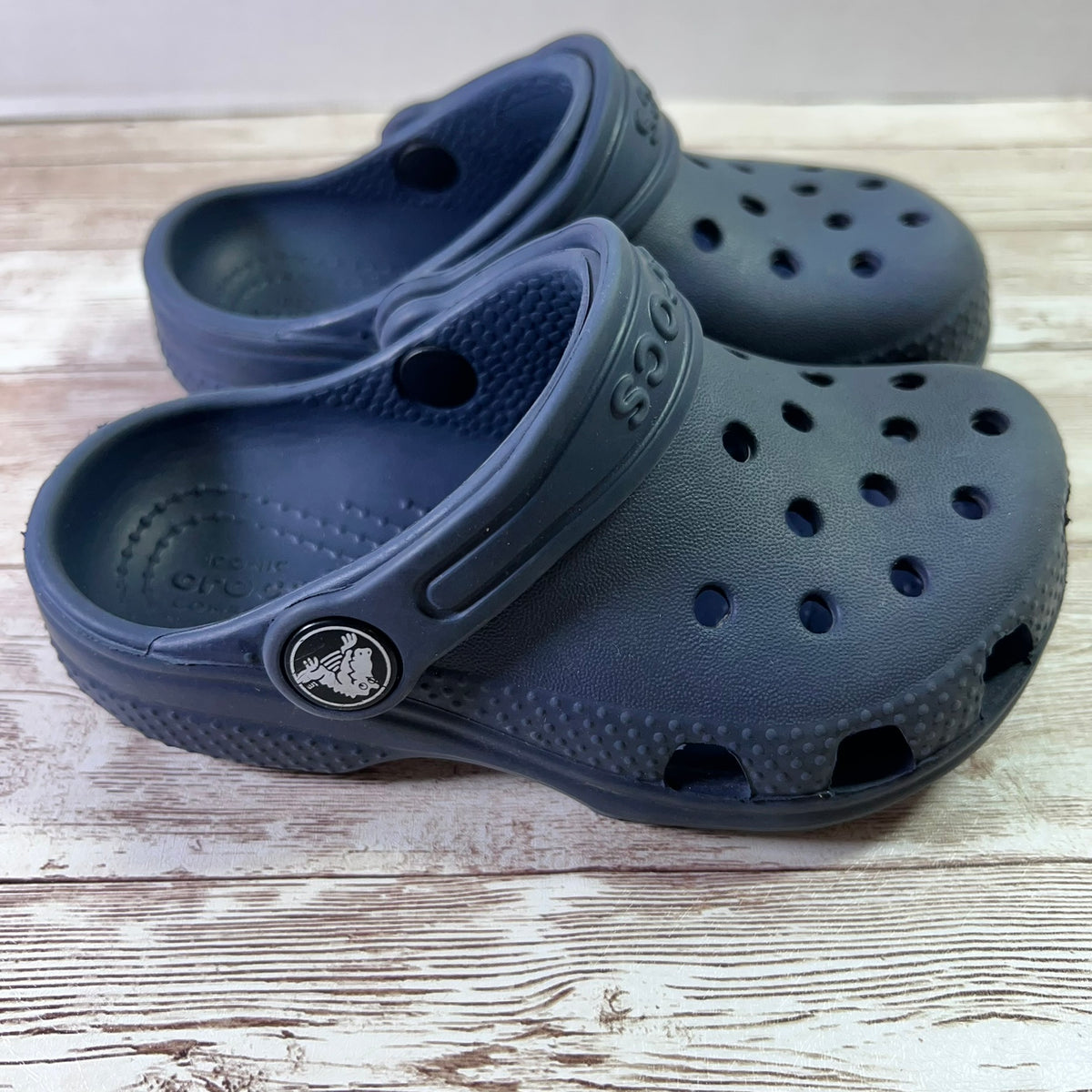 A pair of dark blue Croc-style clogs on a light wood surface, with the brand name partially visible on the strap and a Croc logo charm on the side.