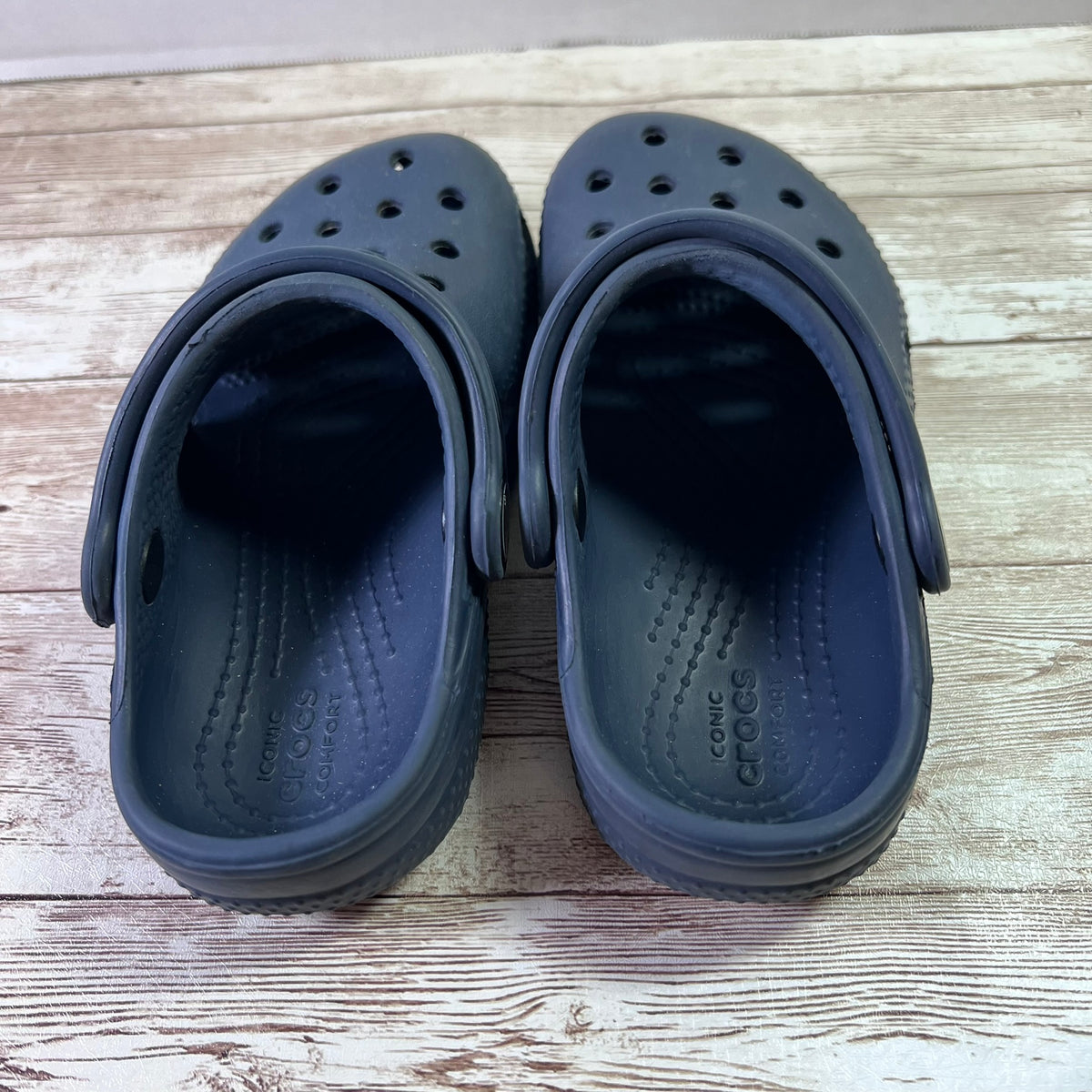 A pair of dark navy Crocs clogs shown from the back, resting on a light-colored wooden surface.