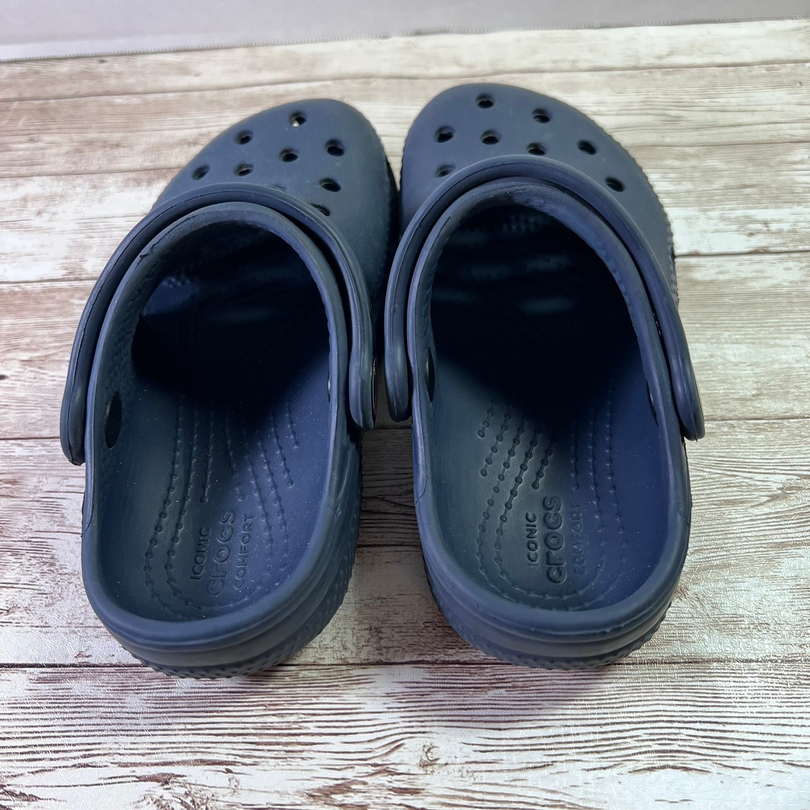 A pair of dark navy Crocs clogs shown from the back, resting on a light-colored wooden surface.