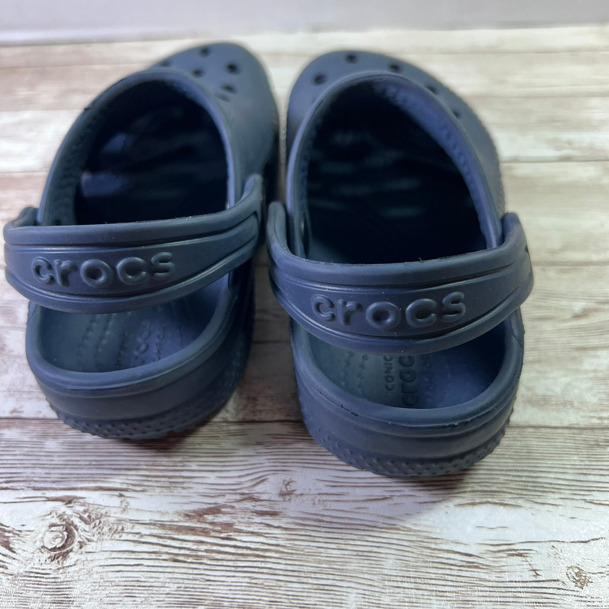 A pair of dark gray Crocs clogs shown from the back on a light-colored wooden surface.