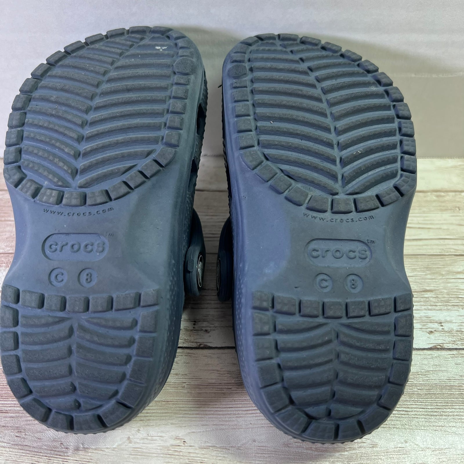 The soles of a pair of dark gray Crocs shoes, showing the textured tread pattern and embossed Crocs branding, photographed from above on a light wood surface.
