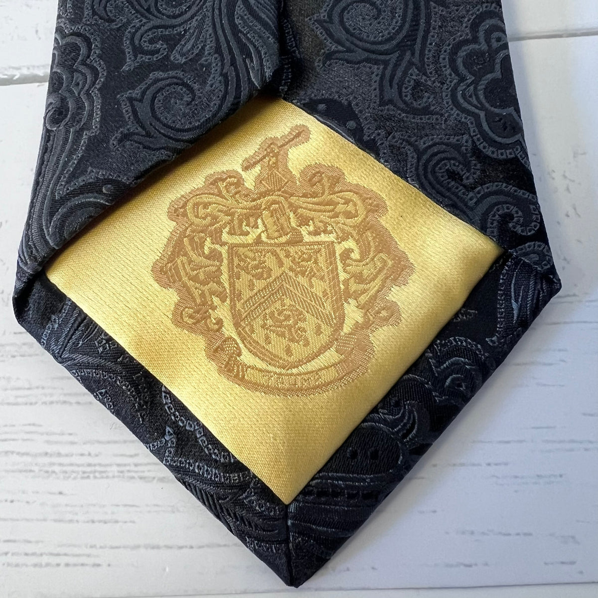 Gold pocket square with a crest design on a black patterned tie.
