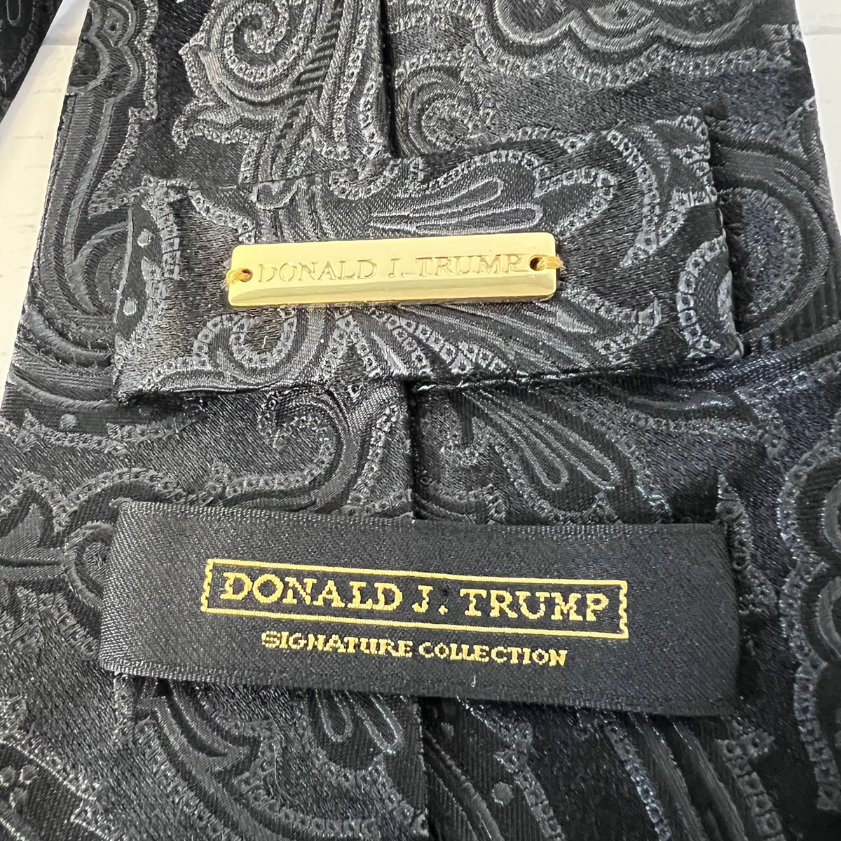Black paisley tie with gold and black label featuring 'Donald J. Trump Signature Collection' on a white background.