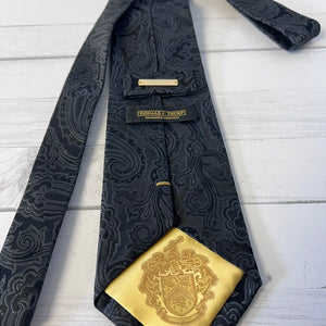 Black paisley tie with gold emblem and label on a light wooden surface