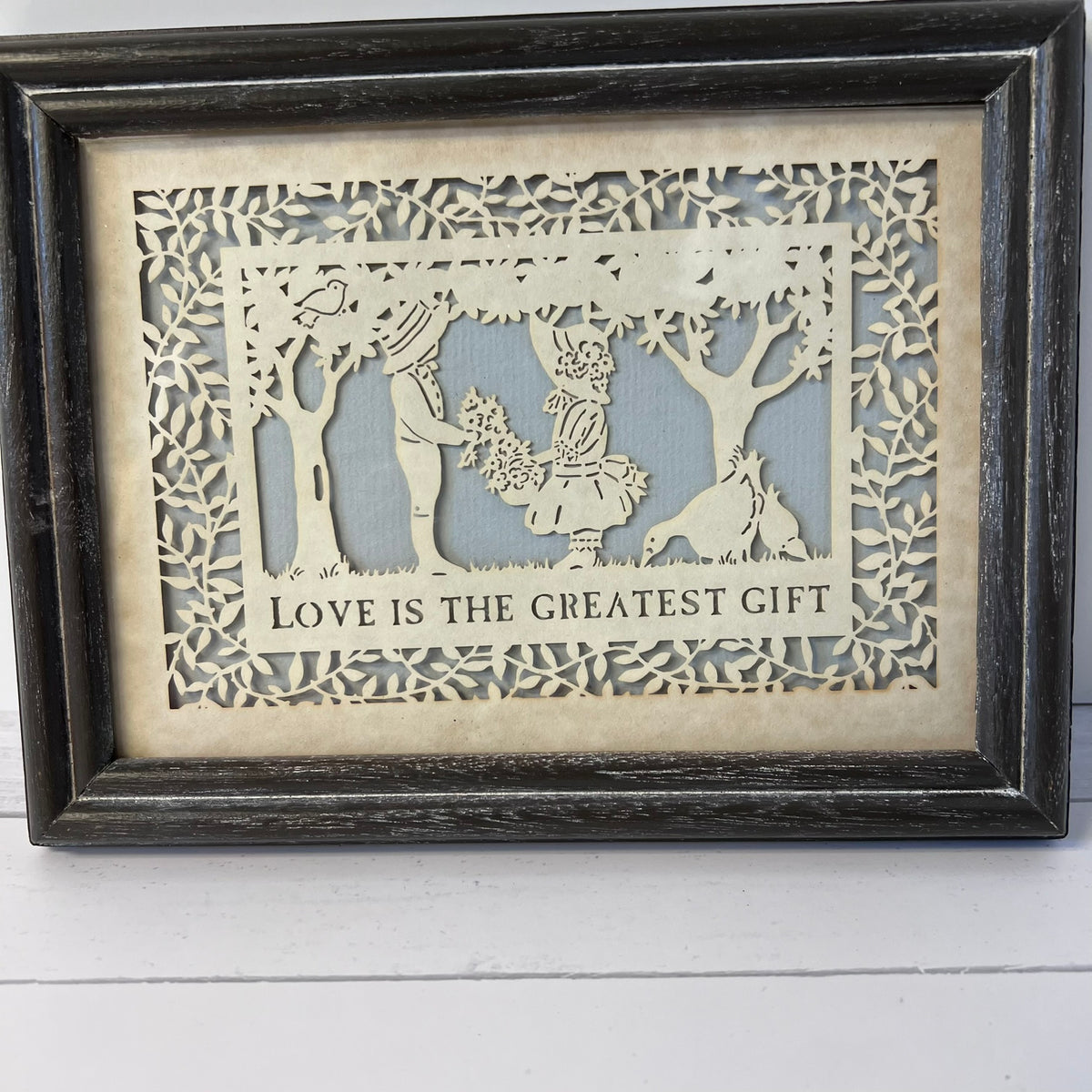 Framed laser-cut wooden wall art with a cutout scene of a couple, trees, and a dog, with the text 'LOVE IS THE GREATEST GIFT' on a light wood background with a dark distressed frame.