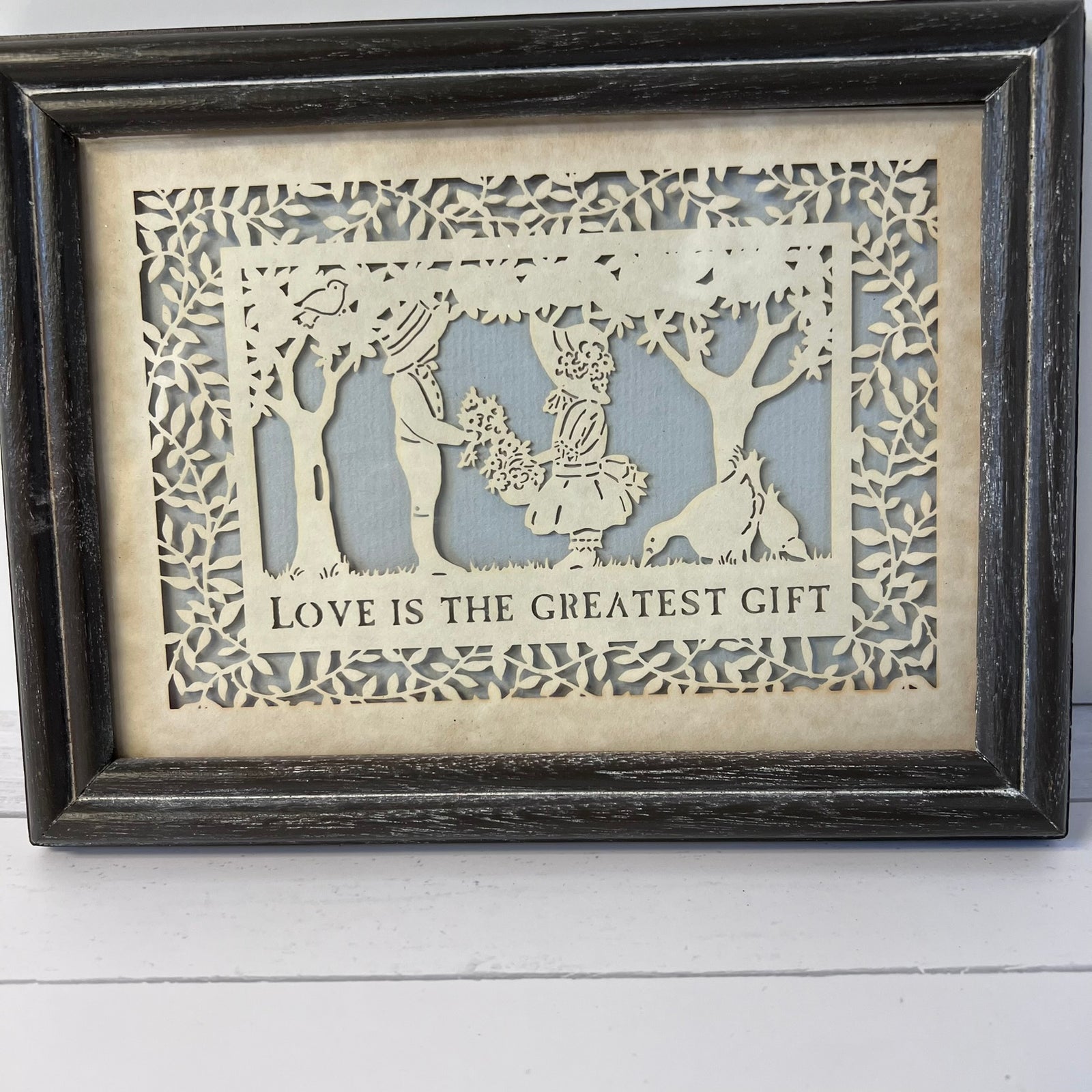Framed laser-cut wooden wall art with a cutout scene of a couple, trees, and a dog, with the text 'LOVE IS THE GREATEST GIFT' on a light wood background with a dark distressed frame.