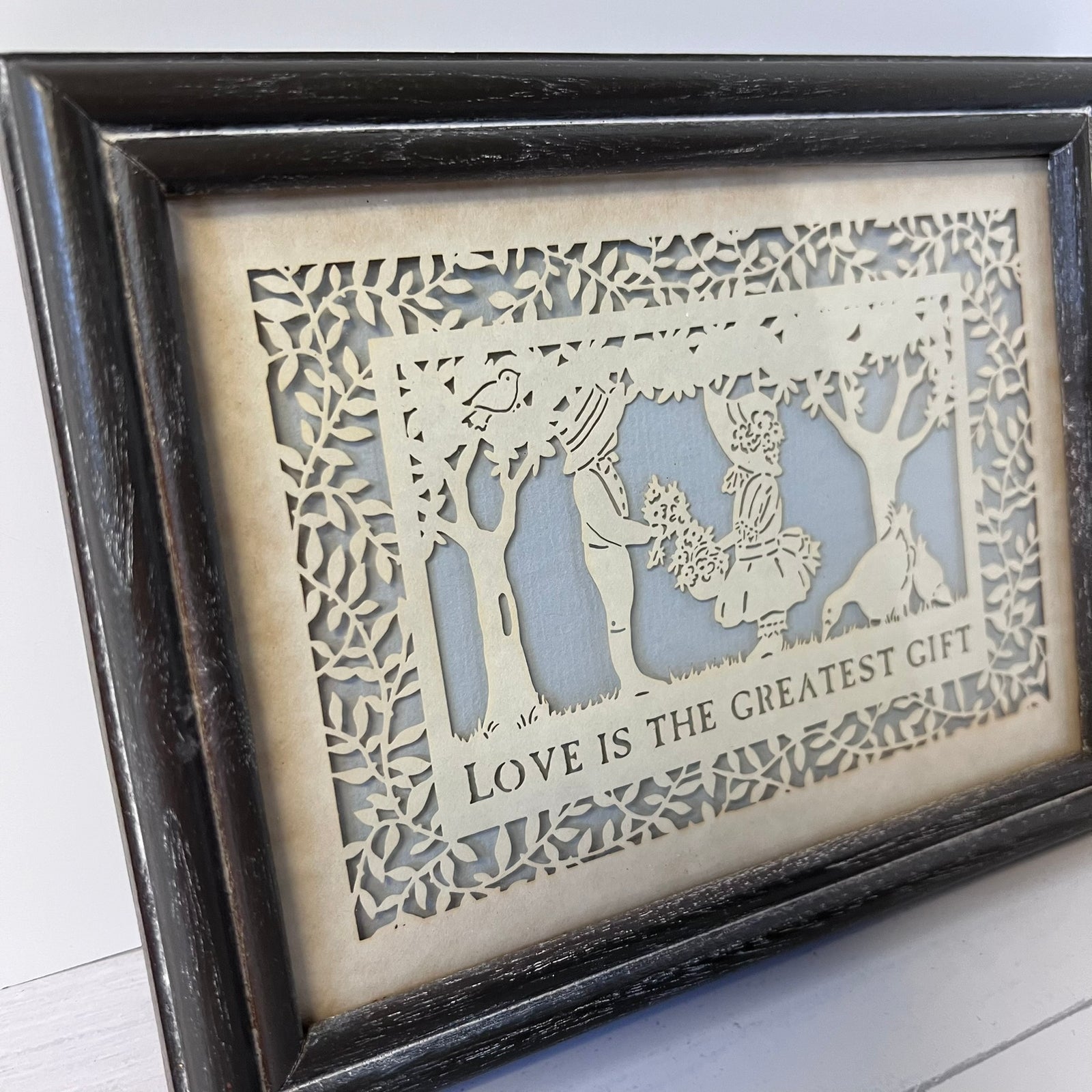 A framed laser-cut wooden art piece featuring a forest scene with a woman and dog, with the text 'LOVE IS THE GREATEST GIFT' at the bottom, set in a dark distressed wood frame.