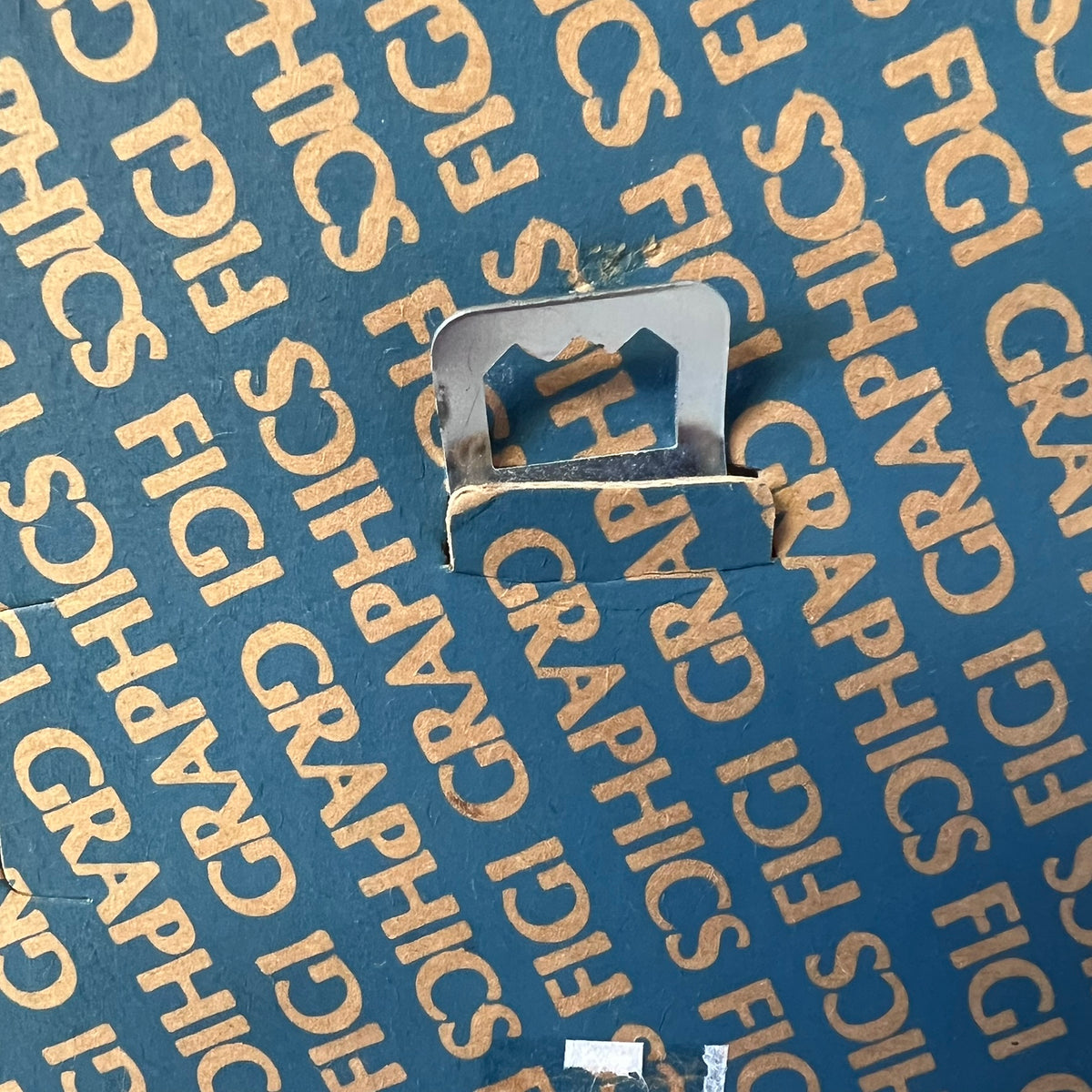 A close-up of a small silver metal clip or fastener placed on blue protective packaging printed repeatedly with the words 'FIGI GRAPHICS'.