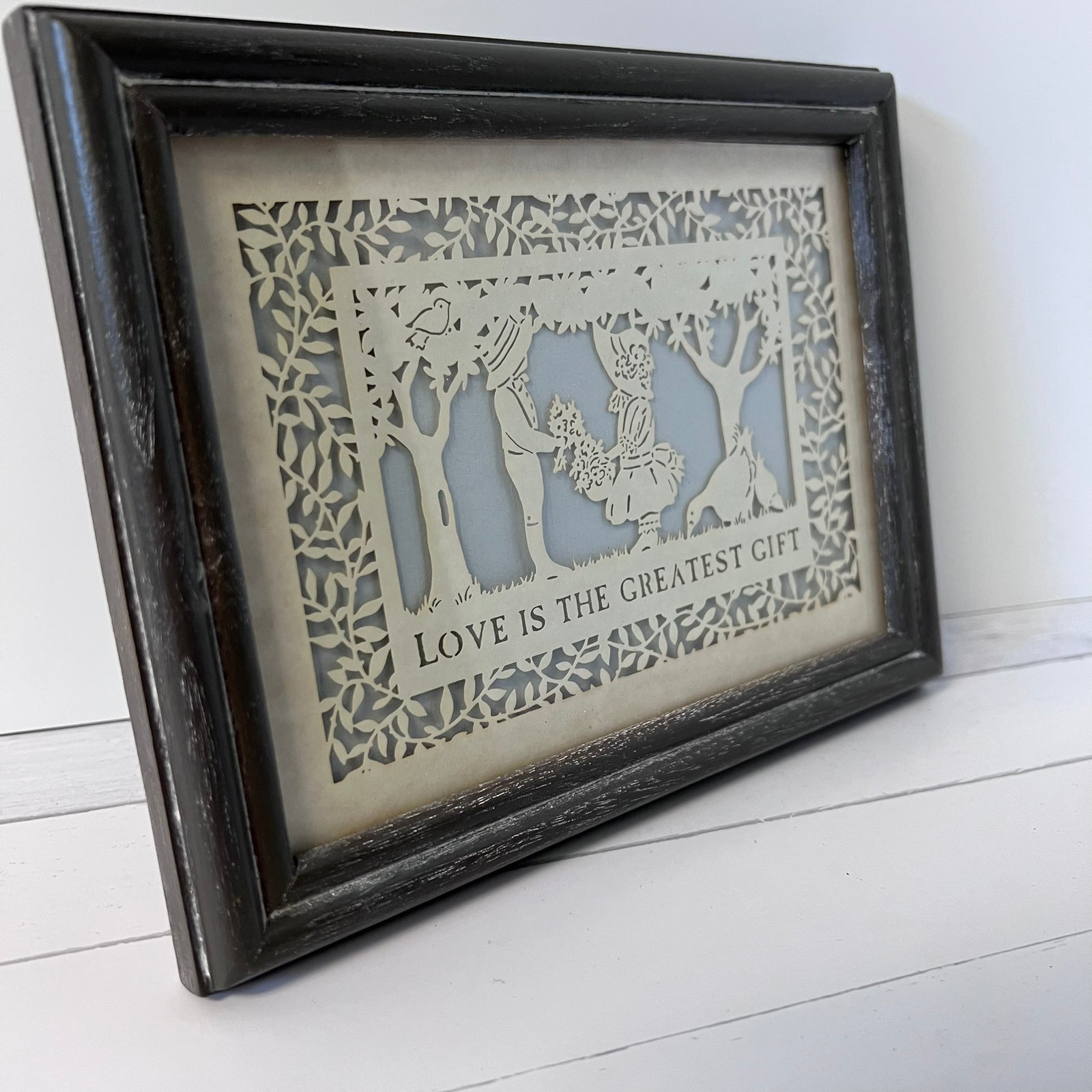 A framed laser-cut paper art piece featuring a couple with a baby and the text 'LOVE IS THE GREATEST GIFT', set in a distressed dark wood frame against a white background.