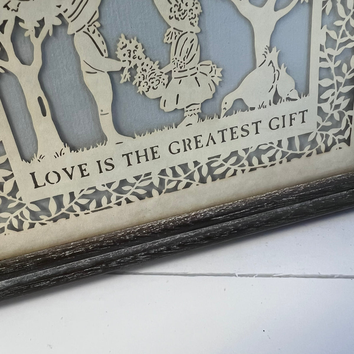 Close-up of a laser-cut wooden decorative piece with layered silhouettes of figures, trees, and a dog, plus the phrase 'LOVE IS THE GREATEST GIFT' on a light wood panel. A dark rustic frame is visible at the bottom.