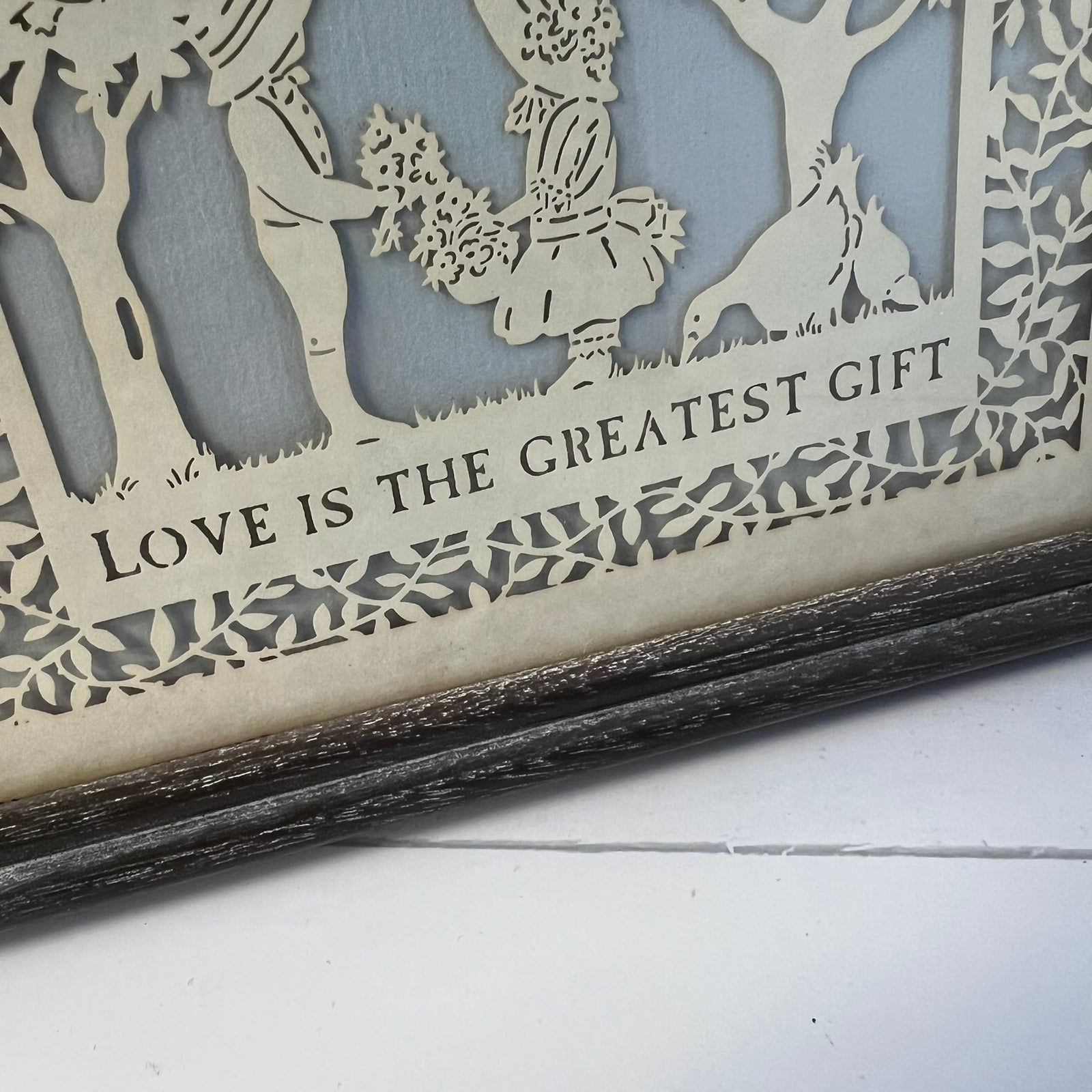 Close-up of a laser-cut wooden decorative piece with layered silhouettes of figures, trees, and a dog, plus the phrase 'LOVE IS THE GREATEST GIFT' on a light wood panel. A dark rustic frame is visible at the bottom.
