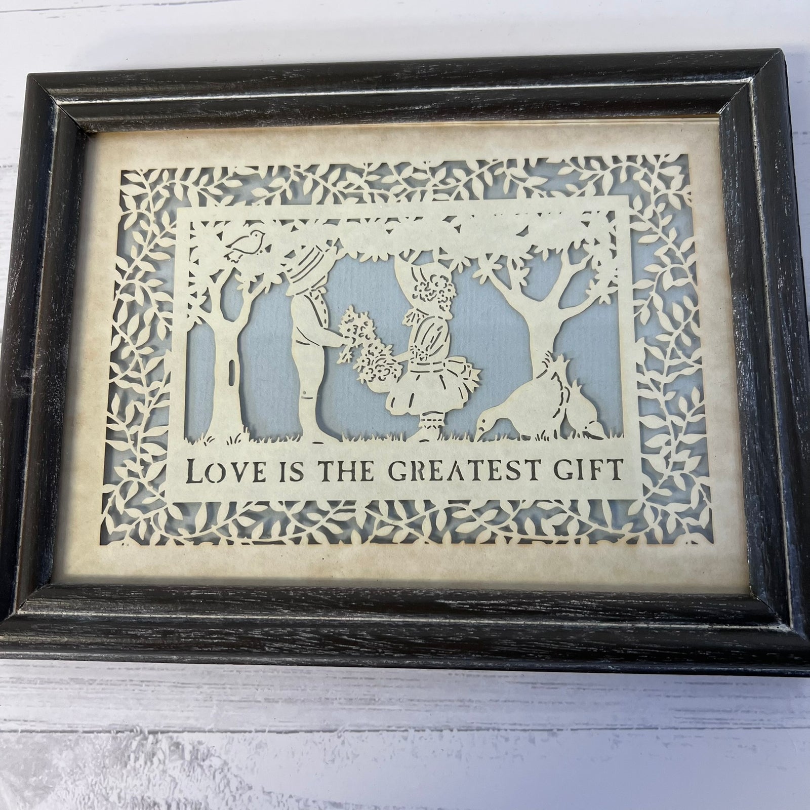 Framed laser-cut wooden wall art featuring a couple and a dog among trees with the text 'LOVE IS THE GREATEST GIFT' on a light wood background with a dark distressed frame.