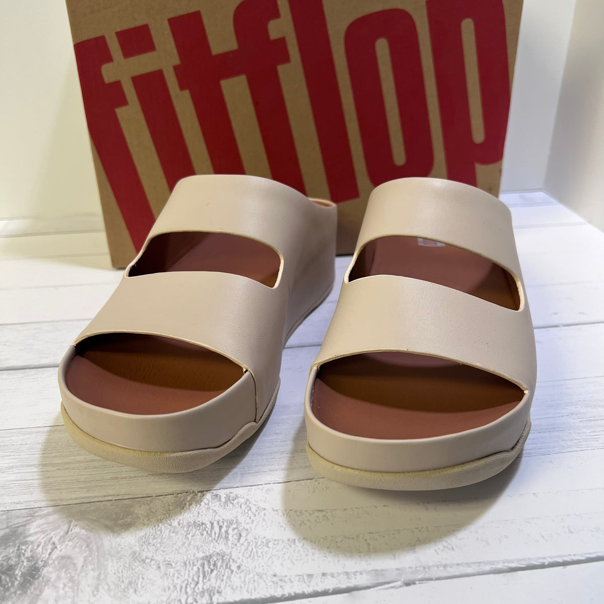 A pair of beige double-strap platform slide sandals with brown footbeds, shown next to their brown and red Fitflop shoebox on a white wooden surface.