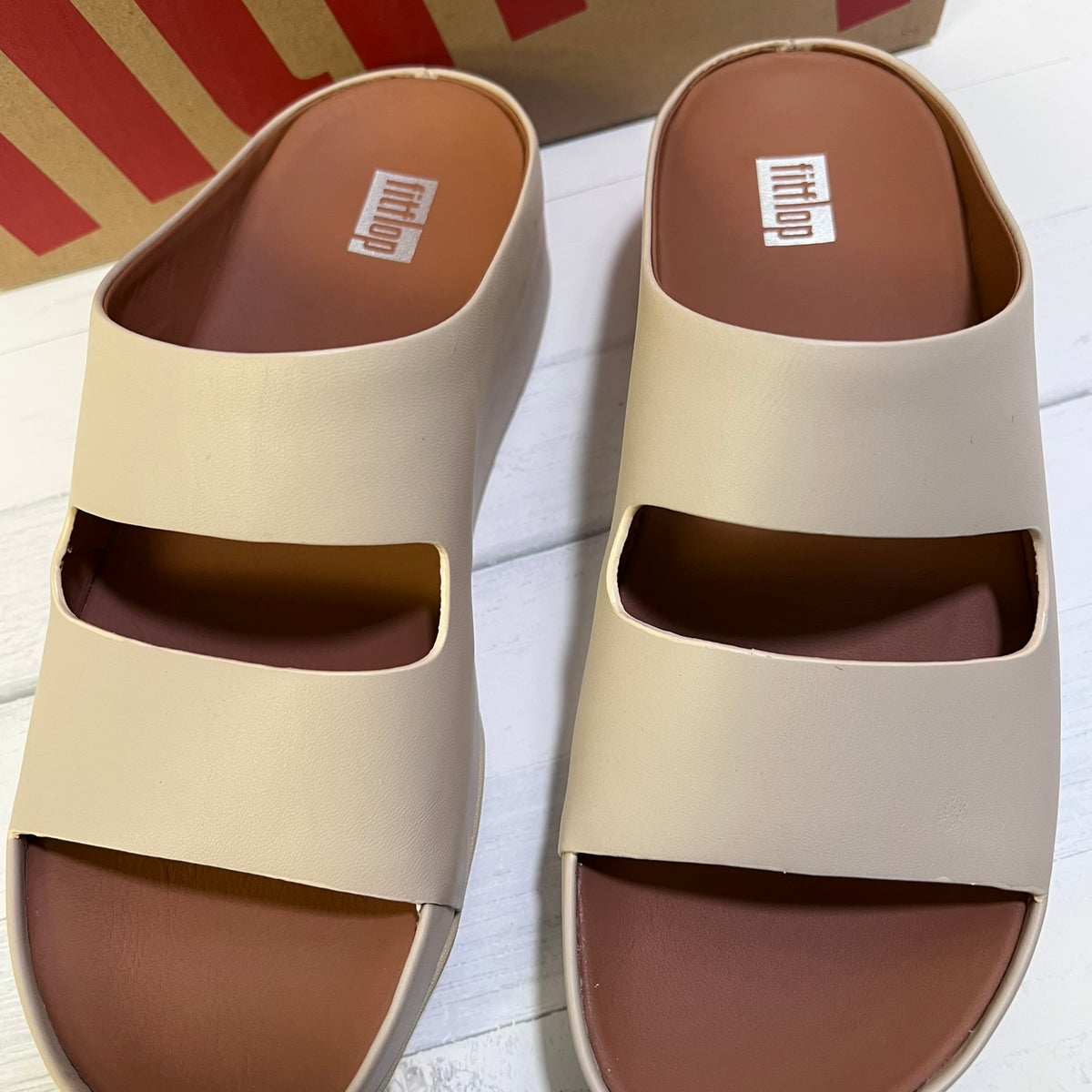 A pair of beige double-strap slide sandals with brown footbeds, shown on a white wood surface with a red and brown shoebox in the background. The brand name 'mittlo' is visible on the insoles.