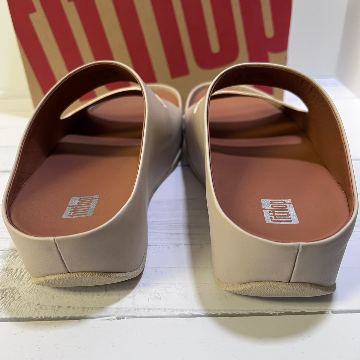 A pair of FitFlop slip-on mule sandals in a light beige color, shown from the back on a white wooden surface with the FitFlop brand box in the background.