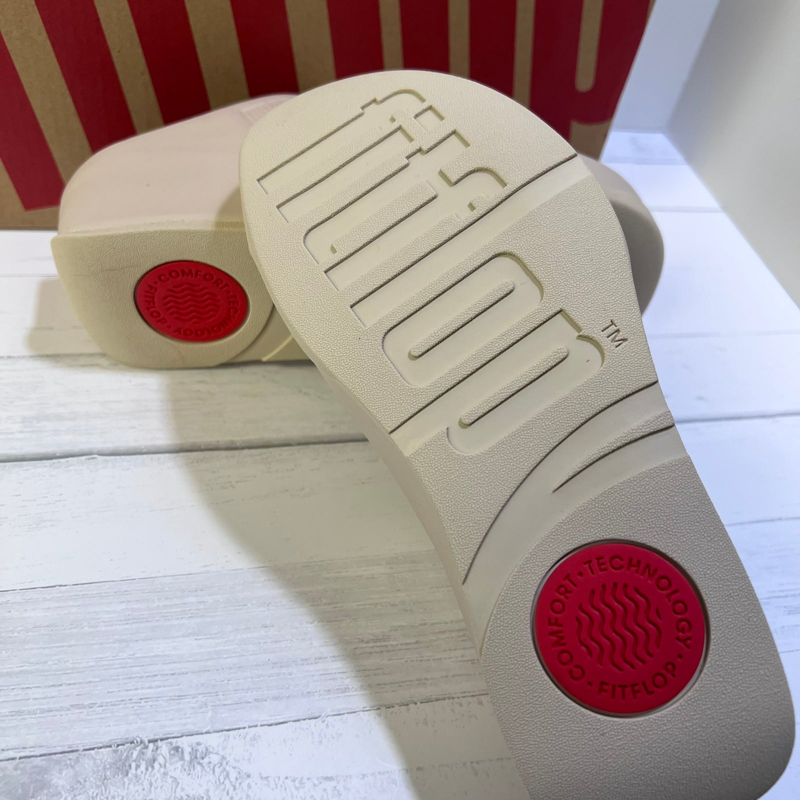 Close-up of the beige sole of a FliPFOE shoe, showing the embossed brand name and a red circular Comfort-Fit logo, with a second shoe and a red-and-beige box in the background.