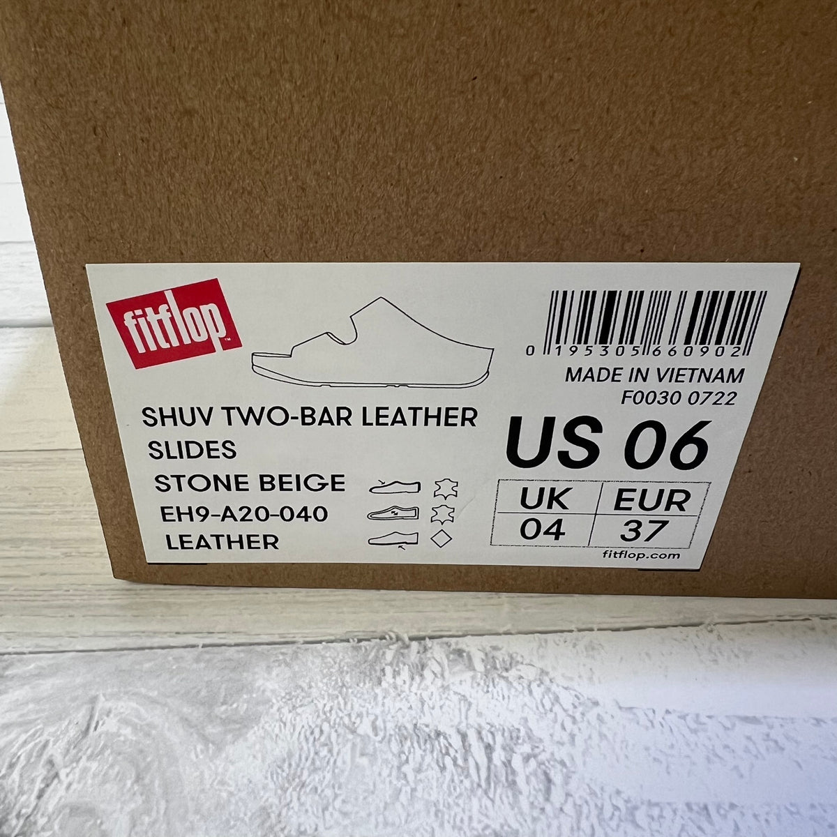 A close-up of a brown cardboard shoe box showing a white product label for fitflop Shuv Two-Bar Leather Slides in Stone Beige, US 06.