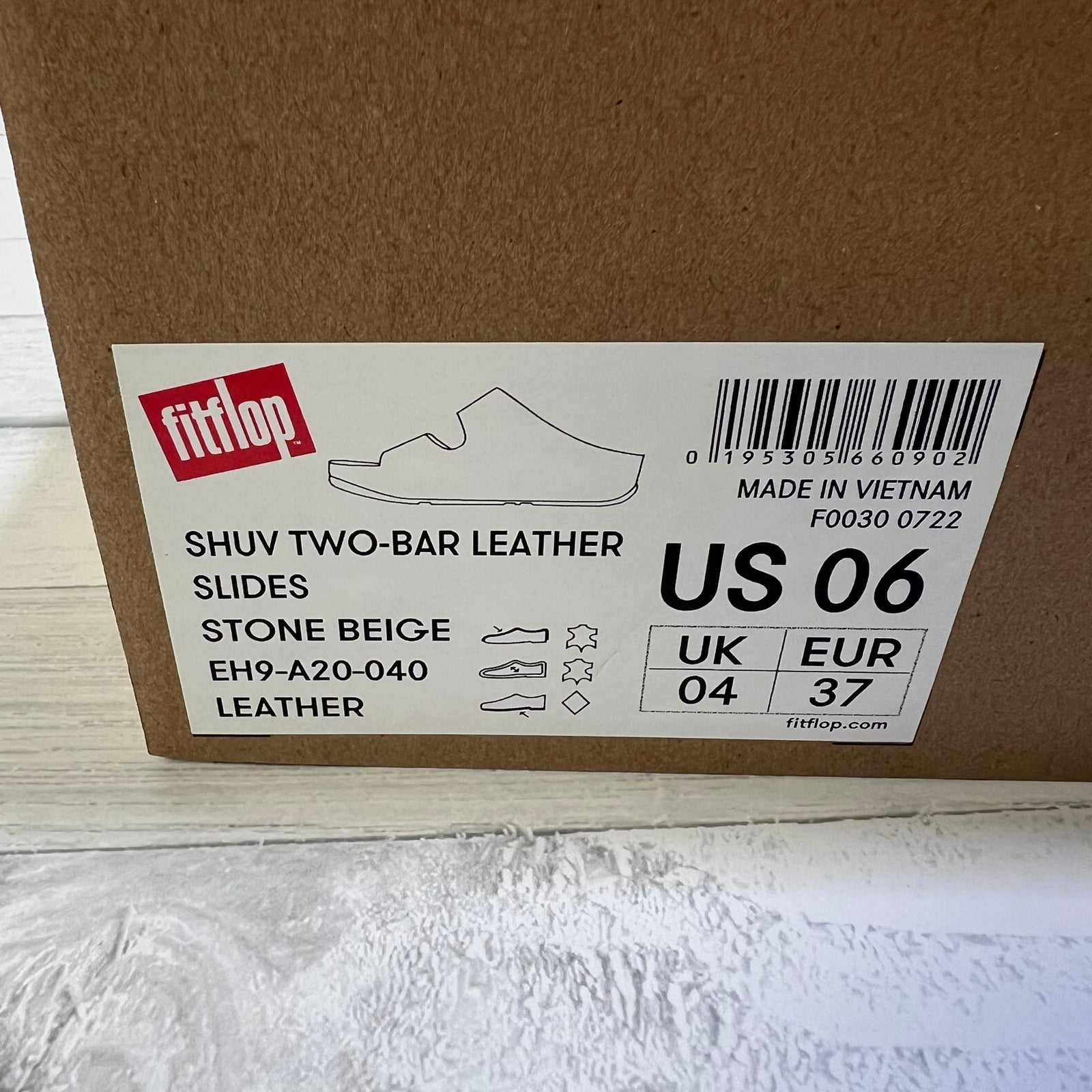 A close-up of a brown cardboard shoe box showing a white product label for fitflop Shuv Two-Bar Leather Slides in Stone Beige, US 06.