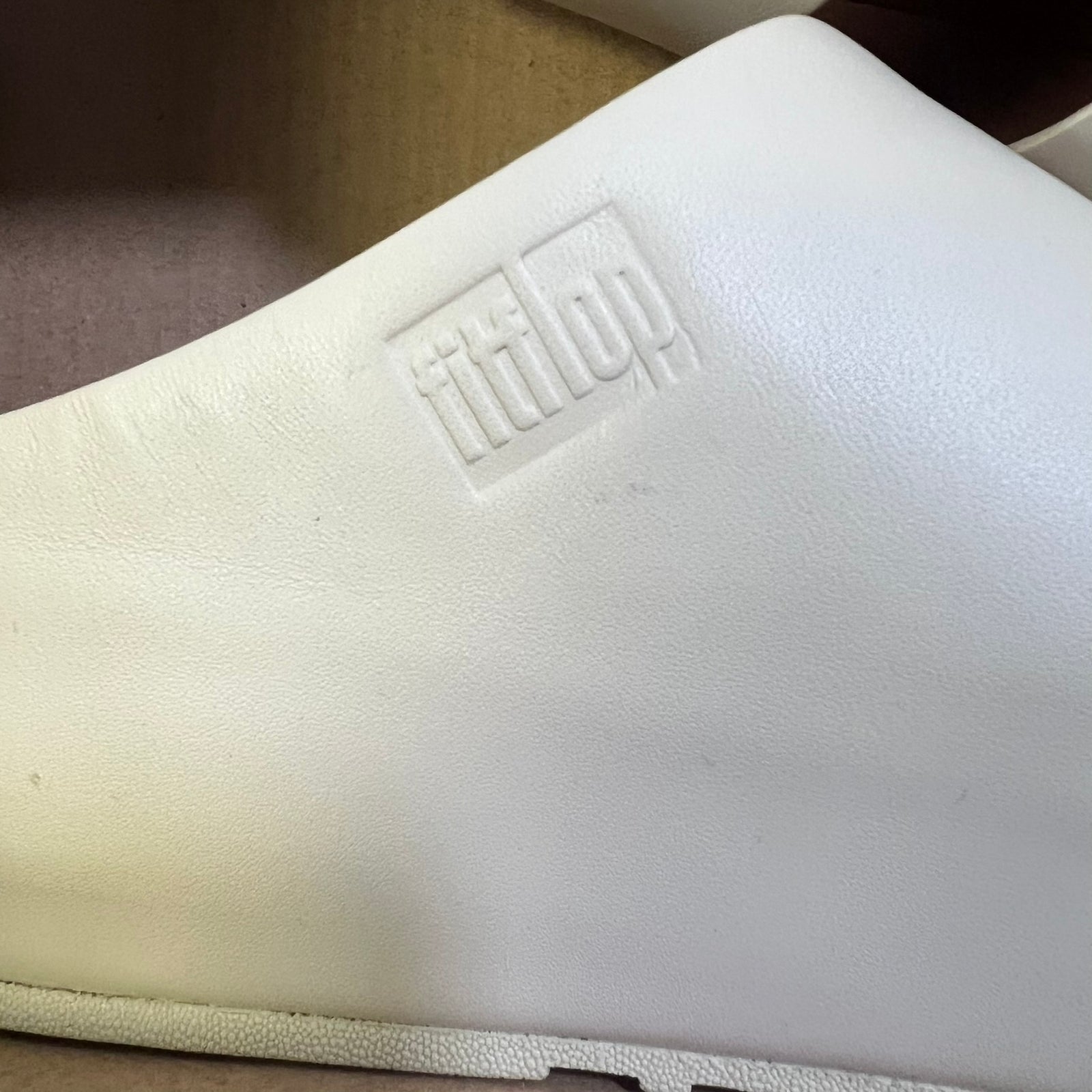 A close-up of a white leather shoe or sandal showing the embossed 'fittop' logo on the side, with a cardboard box interior visible in the background.