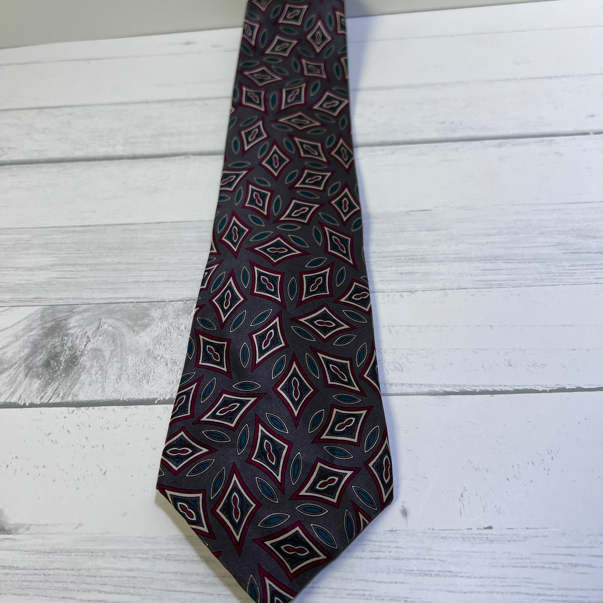 A men's necktie with a dark navy blue base and a repeating geometric diamond pattern in red, white, and teal, laid flat on a white wood plank surface.