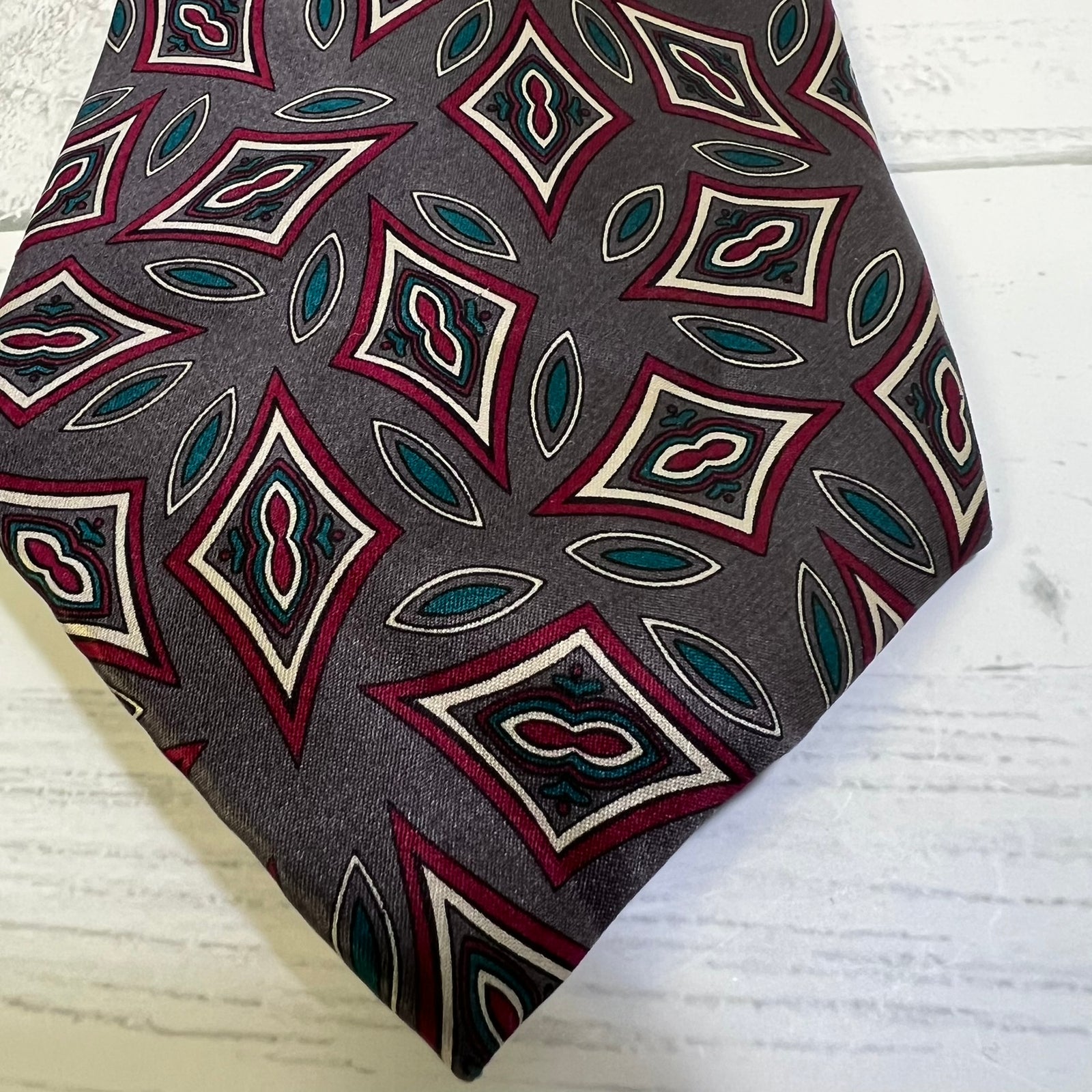A close-up of a patterned necktie with a dark gray base, featuring repeating diamond and leaf motifs in burgundy, cream, and teal, laid flat on a white wood surface.