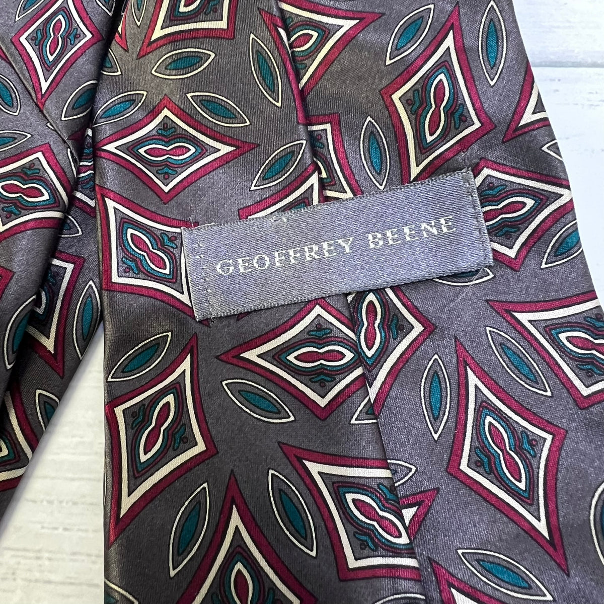 A close-up of a Geoffrey Beene patterned necktie in gray, featuring red, teal, and cream geometric diamond motifs, with the brand name clearly visible on the woven label.