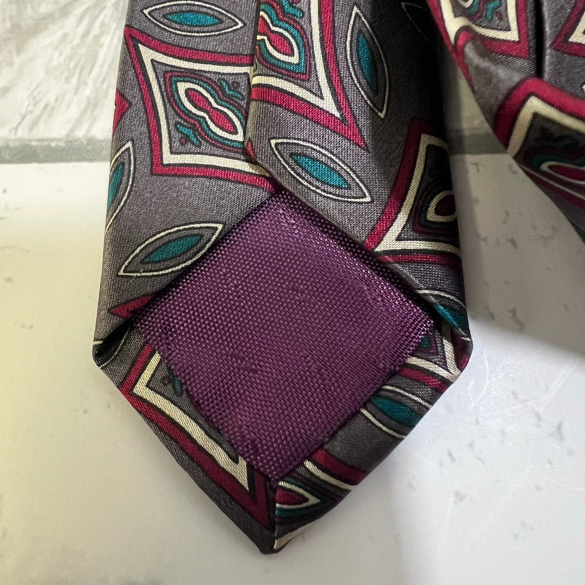 A close-up of a patterned necktie with a gray base, featuring geometric diamond motifs in teal, burgundy, and cream, with a solid purple fabric band at the tip.