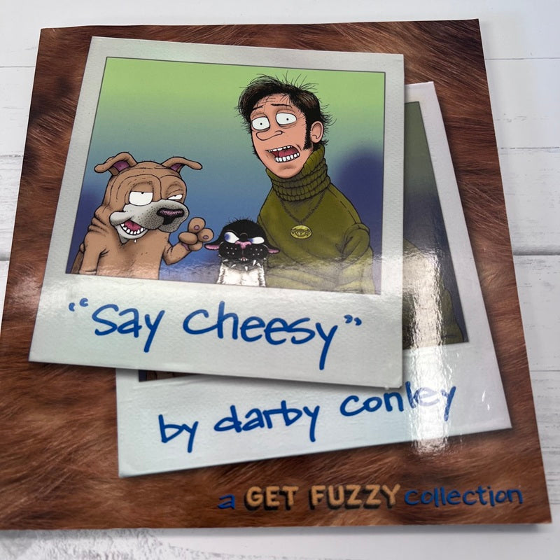 Cartoon illustration of a dog and a person with text 'Say cheesy' by Darby Conley on a wooden surface.