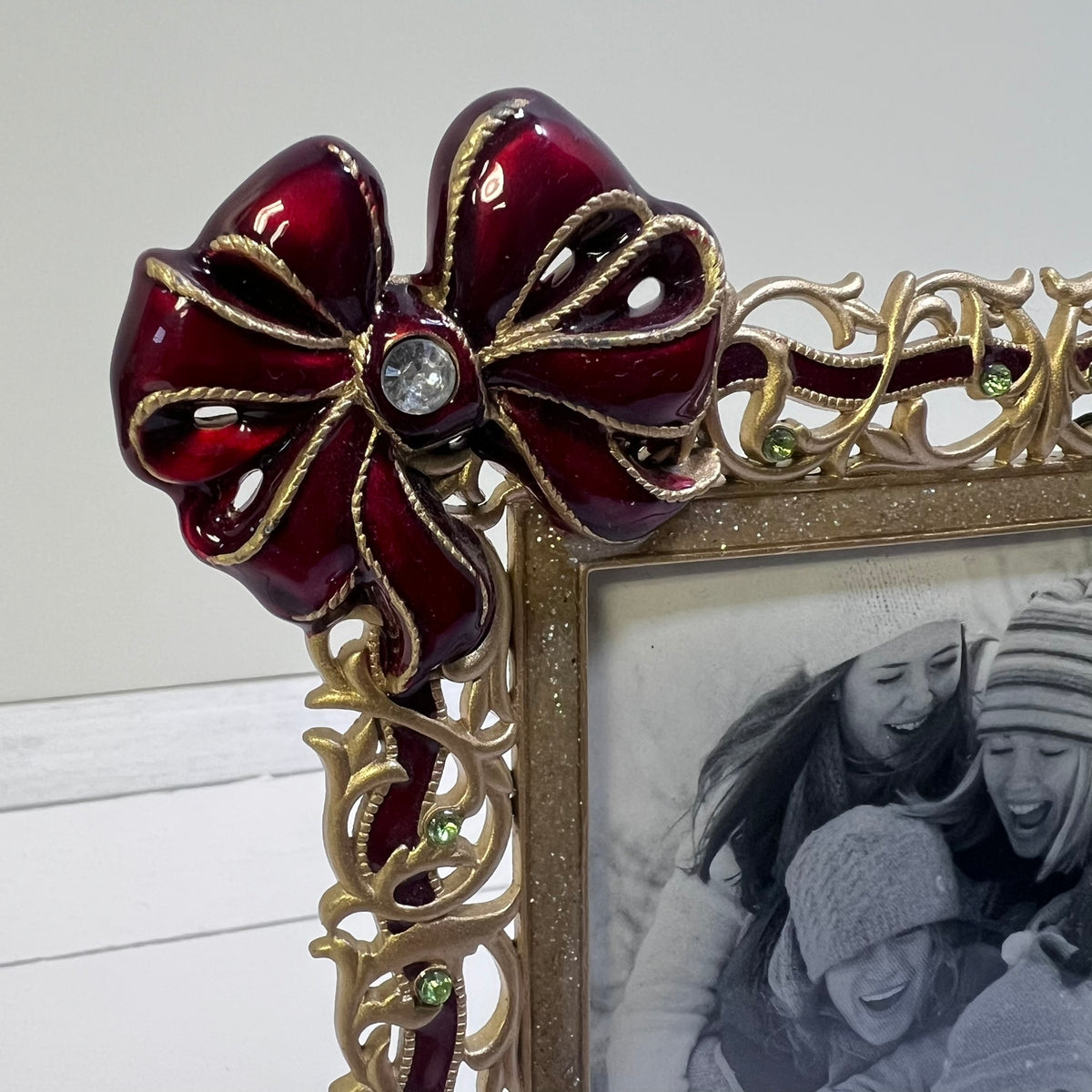 A close-up of an ornate picture frame corner featuring a glossy deep red bow embellishment with gold trim and a clear rhinestone center, alongside a black-and-white family photo insert.