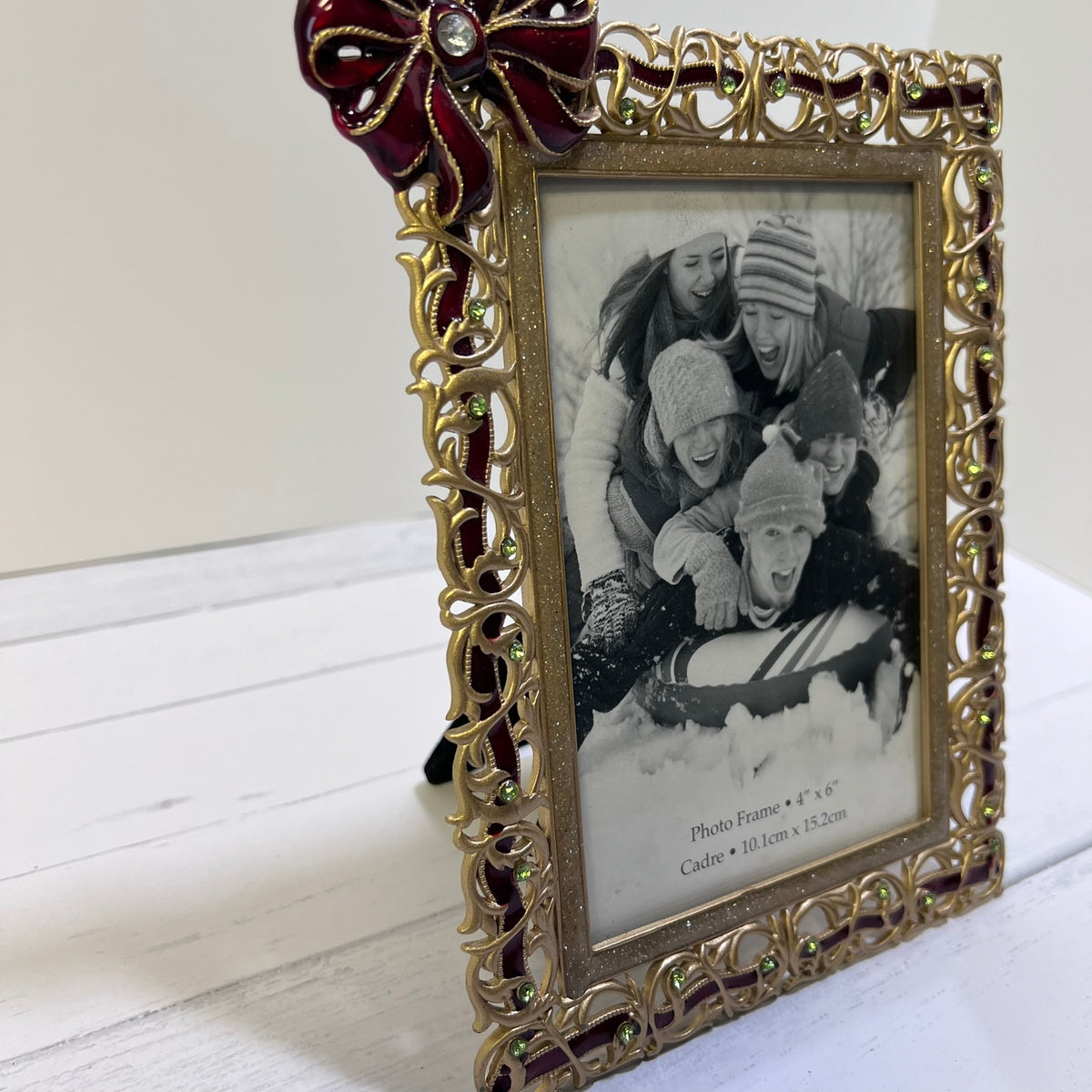 An ornate gold picture frame with a decorative red bow on the top left corner, holding a black-and-white family photo. The frame has intricate scrollwork and small green crystal accents, standing on a white wooden surface against a light background.