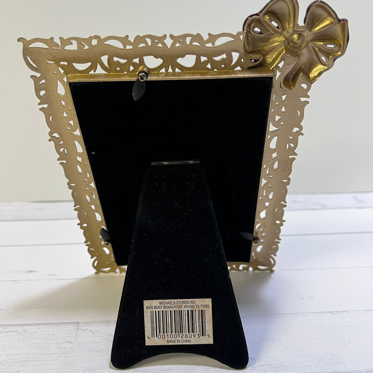 A decorative gold picture frame with an ornate filigree border and a bow detail on the top right corner, shown standing on a black velvet display stand against a white background.